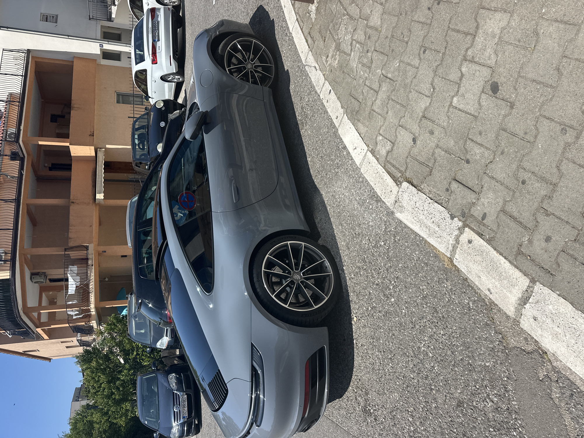 The image shows a grey Porsche 911 parked on a street in Budva, Montenegro. The car is facing right, with its rear and side visible. The street is paved with asphalt and a sidewalk made of interlocking concrete pavers is visible on the right side of the image. In the background, there is a building with multiple balconies and trees. Other cars, including a white Audi and a dark Volkswagen, are also parked on the street. The sky is clear and blue, indicating a sunny day.