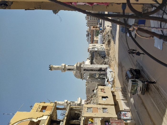 The image depicts a devastated urban street scene in Khan Yunis, Palestinian Territory, viewed from an elevated position. Dominating the midground is a severely damaged mosque with a tall, slender minaret, featuring small turrets at its apex. The mosque's main dome and surrounding structures are largely reduced to rubble, which piles extensively around its base. To the left, a multi-story building, appearing residential, exhibits significant structural damage with exposed interiors, shattered windows, and large sections of facade missing; laundry hangs from various balconies. An ornate railing from the observer's building frames the right foreground. The paved street below is dirty and covered with scattered debris. A white tanker truck, marked "INTER 4509," is visible on the left side of the street, facing left. Six individuals are walking on the street: one person is crossing in front of the truck, and a group of five are further down the street on the right, walking away from the camera. Two parked cars are also visible further down the street on the right. The background shows more urban buildings, some appearing damaged, beneath a clear blue sky.