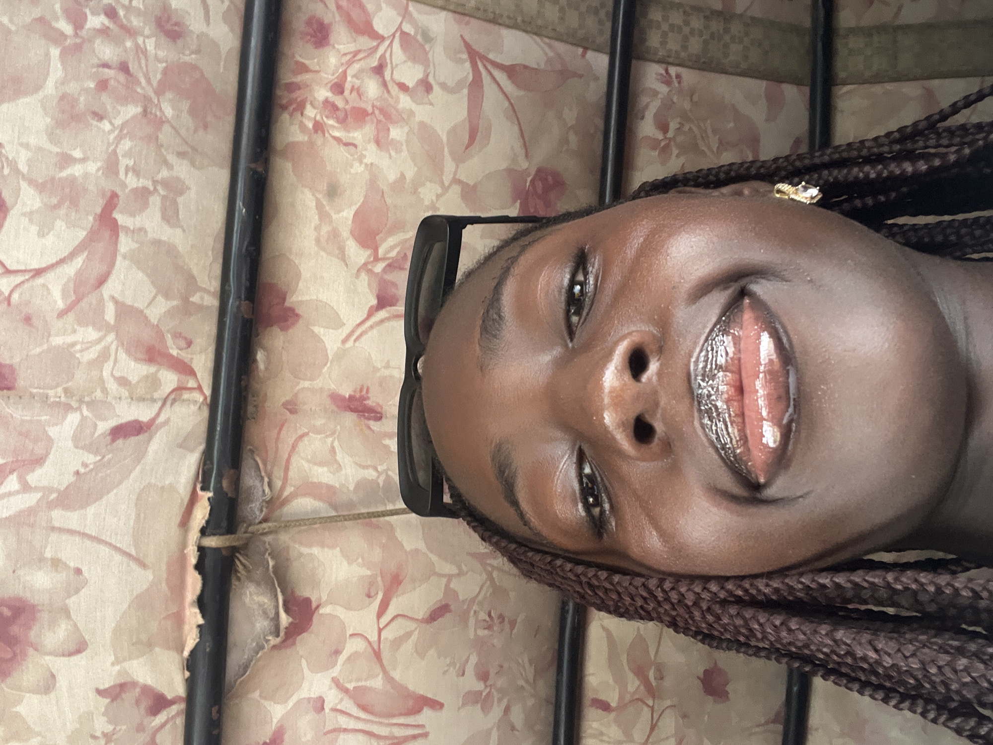 The image, oriented on its side, presents a close-up portrait of a young Black woman from Karum, Nigeria, smiling towards the camera. She has rich brown skin and braided hair, which cascades along the right side of the frame. Black sunglasses rest on top of her head, and a small, gold-toned earring with a clear stone is visible on her upper right ear. Her lips are accentuated with a glossy, light brown or metallic finish.
The background features a light, possibly beige, surface adorned with a repeating floral pattern in shades of pink and muted red. A dark, thin metal bar runs vertically through the left side of the frame, showing signs of wear and tear, and is secured by a piece of twine. The woman appears to be posing for a casual photo, with an expression of happiness. No specific time of day or weather conditions are discernible from the lighting, and there is no visible text.