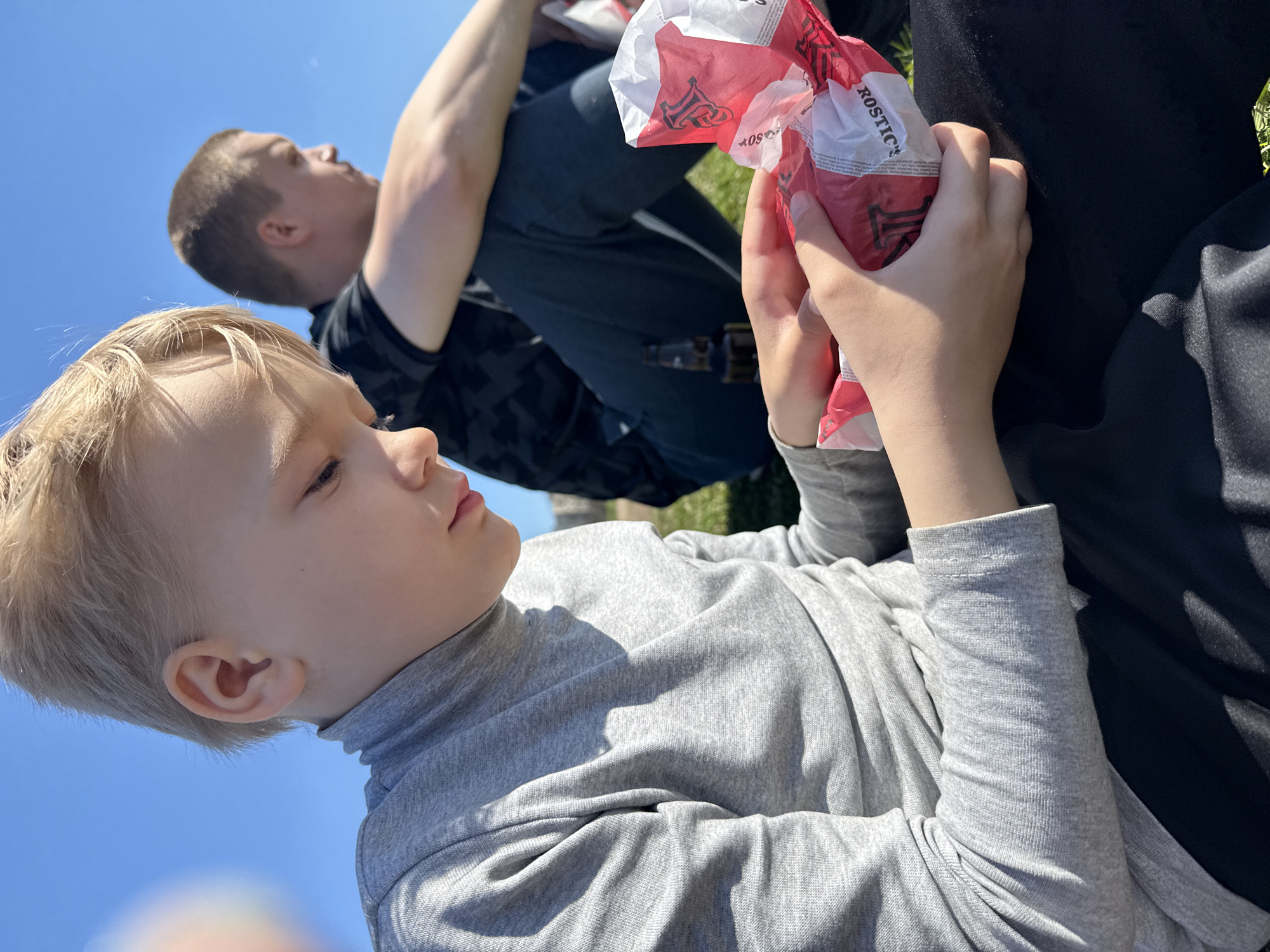 Two boys are outside on a sunny day. A younger boy with blond hair is lying on his side and looking at a red and white paper bag that he holds in his hands. The bag says "ROSTIC'S" on it. An older boy is behind him, sitting with his arms crossed. The background is blue sky. The location is Dmitrov, Russia.