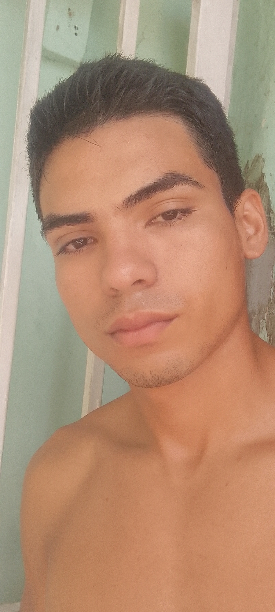 A close-up head and shoulders shot of a light-skinned young man with dark hair and eyebrows. He has brown eyes and a hint of stubble on his chin. He is shirtless. Behind him, there is a white painted structure consisting of vertical and horizontal beams. The background appears to be a light green painted wall. The location is claimed to be Juan Griego, Venezuela, but there are no visual elements confirming the city in the image.