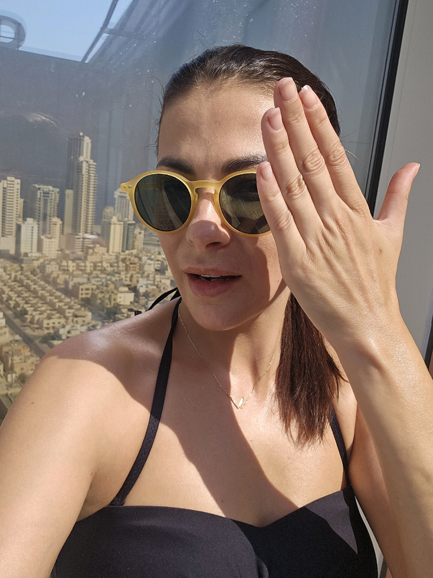 A woman is positioned in the foreground of the image, partially obscuring her face with her right hand. She is wearing yellow-rimmed sunglasses, a black bikini top, and a delicate gold necklace with a small pendant. Her hair is pulled back, and her skin appears to be glistening, possibly from sweat or sunscreen. The background reveals a panoramic view of a city skyline, identifiable as Manama, Bahrain, with numerous high-rise buildings and a hazy, urban landscape under a bright sky. The lighting suggests it is daytime, with strong sunlight casting shadows.