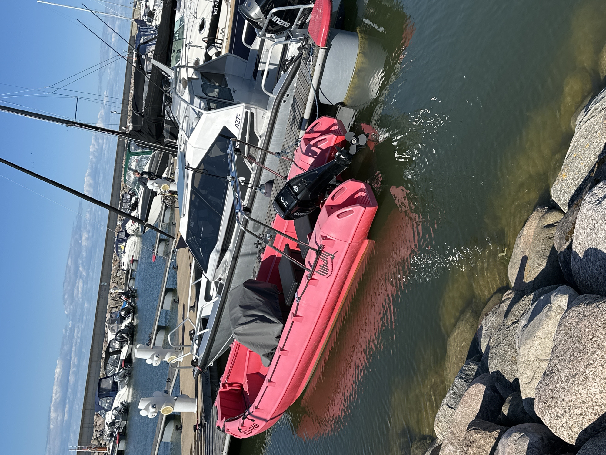 The image depicts a sunny day at a marina in Tallinn, Estonia. In the foreground, a bright pink inflatable boat with a gray cover is docked. The boat has the registration number "YLU-049" and the brand name "Whaly" visible. Behind it, a gray motorboat with a white cabin is also docked, bearing the text "A27c". Several other boats, including sailboats and smaller motorboats, are visible in the background, moored in the marina. A concrete seawall and some rocky shorelines are present. The sky is blue with scattered clouds, suggesting fair weather conditions. No people are actively engaged in any activities in the frame.