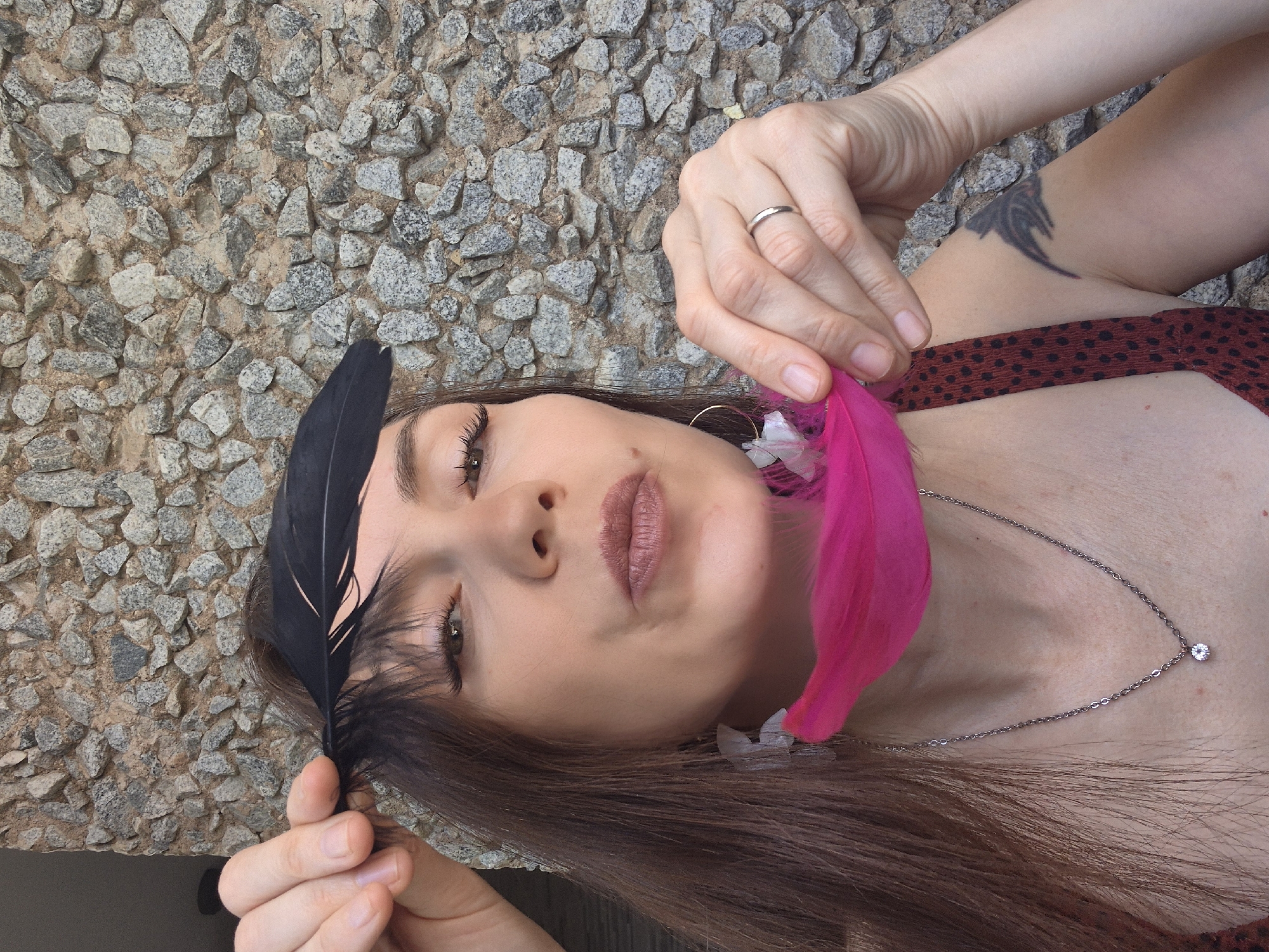 A woman is in front of a stone wall. She has long brown hair, and is holding a black feather above her head with her left hand. She is also holding a bright pink feather with her right hand near her chin. She is wearing a necklace with a diamond pendant and a red and black polka dot top. A tattoo is visible on her upper left arm. The woman's mouth is slightly open, and she appears to be looking at the camera. The image does not provide any specific details or context that would allow for identification of the location in the city of Chaikovsky, Russia.