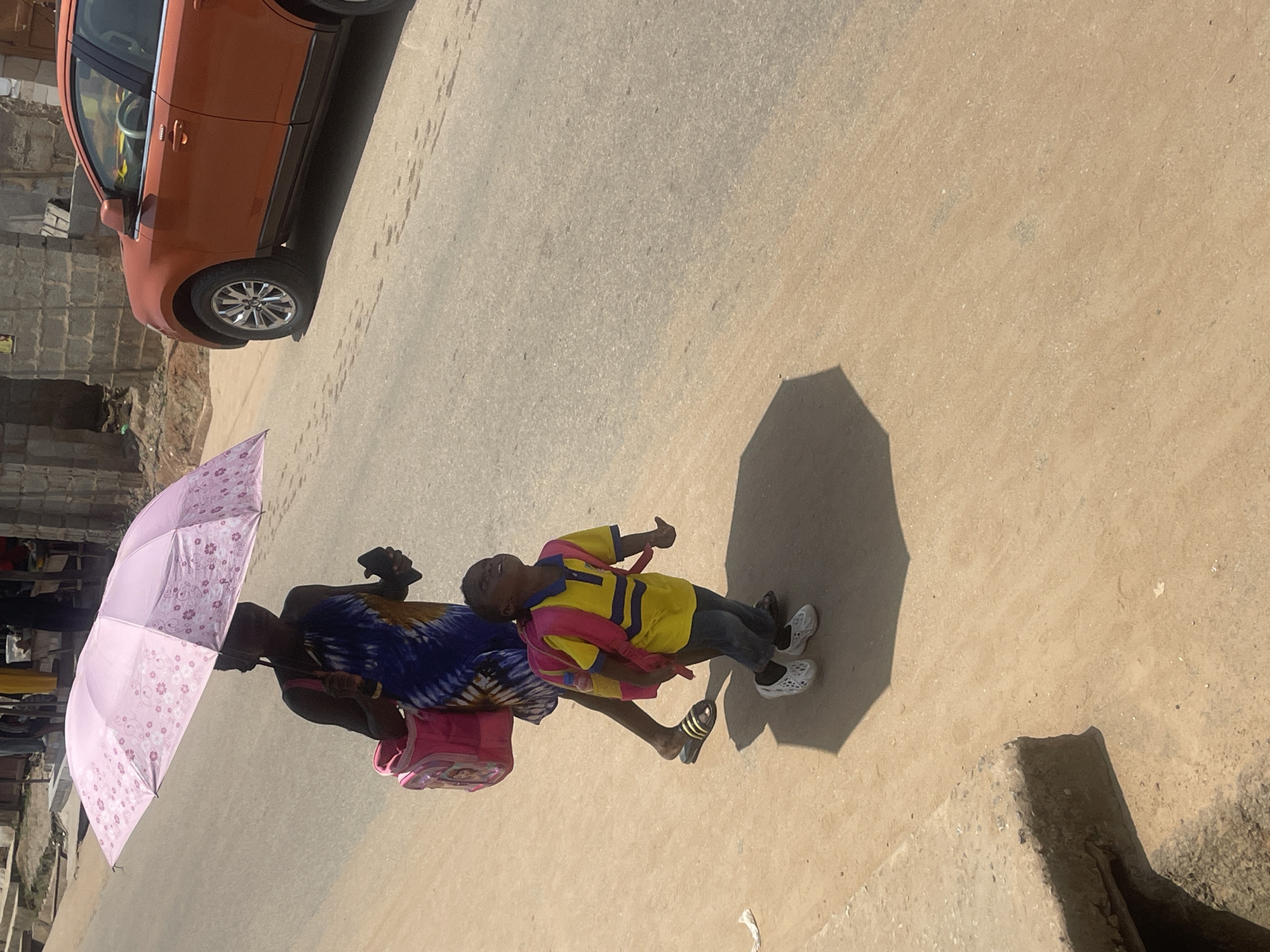 In Abuja, Nigeria, on a dusty, sun-drenched street, a woman walks under a pink floral umbrella, holding a pink child's backpack. Beside her, a young child with a bright yellow shirt and pink backpack looks upwards, seemingly towards the sky. The child wears dark pants and white shoes. Behind them, an orange SUV is parked on the street. The environment is characterized by unpaved ground, a concrete edge on the left, and rudimentary buildings in the background. The scene suggests a warm, possibly hot, day.