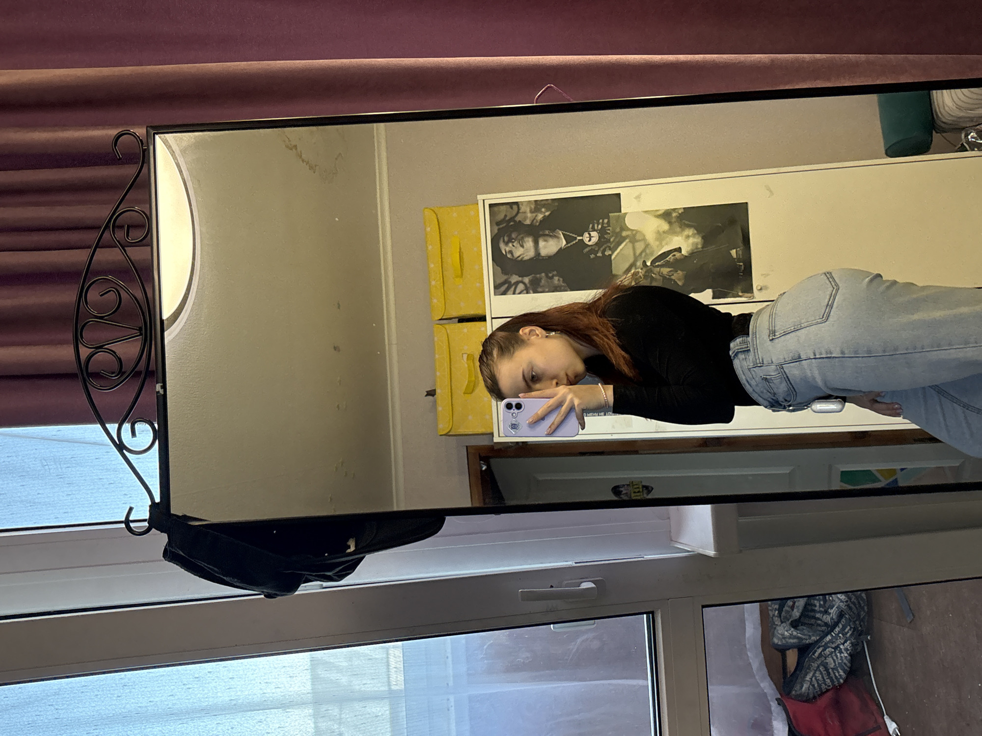 A girl takes a selfie in a mirror. The mirror is hanging in front of a window with a black bag hanging on it. A curtain hangs behind the mirror. Reflected in the mirror is the upper half of a person, a poster, and a storage unit. The location of the city is Прикамский, Russia.