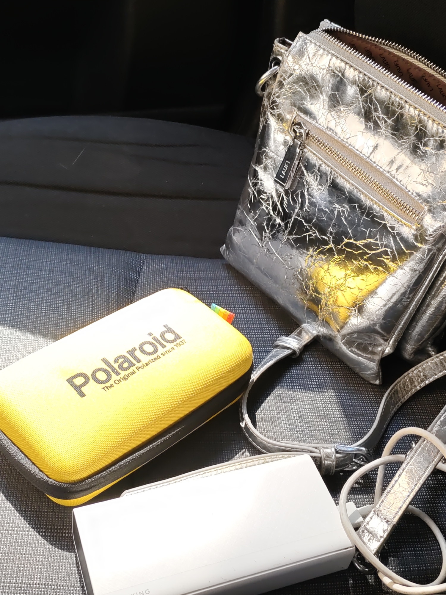 In Rostov-on-Don, Russia, an overhead shot captures a collection of items laid out on a textured gray car seat. A crinkled, silver metallic handbag with a prominent "LAUF!" zipper tag sits to the right. To the left, a bright yellow rectangular case displays the "Polaroid" logo. Below these, a white rectangular object, possibly a power bank, is partially visible, with a white charging cable coiled nearby. The background is a dark car interior, creating a stark contrast with the brightly lit objects.