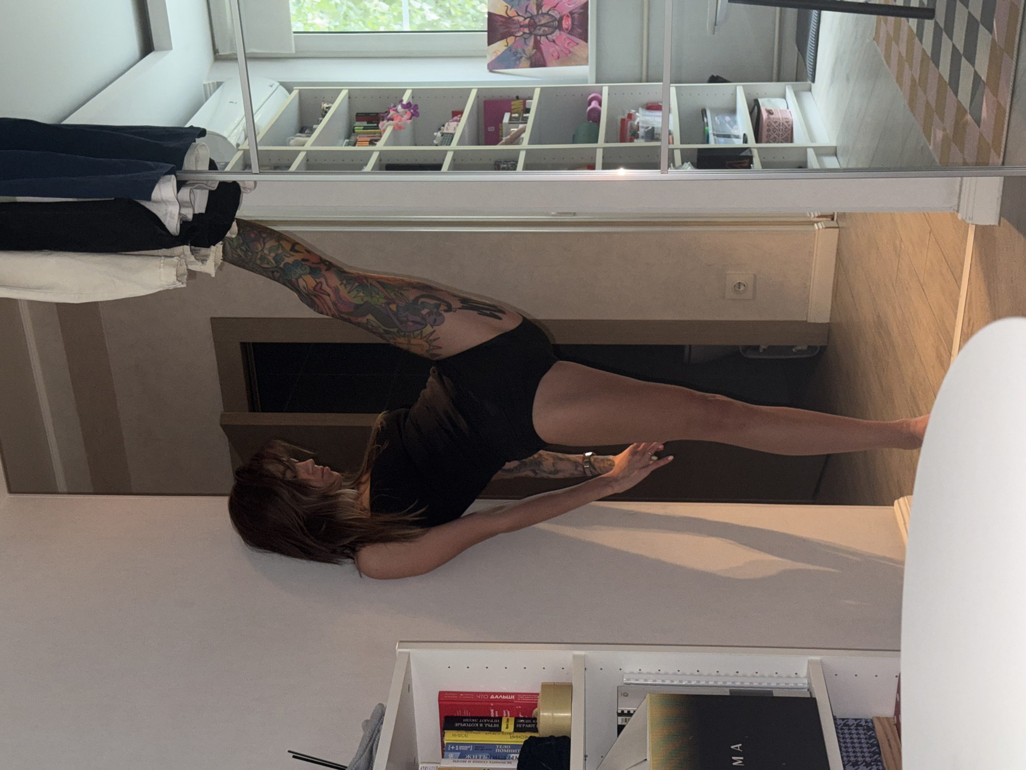 A woman with long brown hair is performing a split on a dark mat. She is wearing a black tank top and black shorts. Her body is covered in colorful tattoos. She is in a room with white shelves filled with books and decorative items. A window is visible in the background, showing trees and a blue sky. The room appears to be well-lit, suggesting it is daytime. The overall mood is one of flexibility and athleticism. On the shelves, several books with Russian titles are visible, including "ЧТО ДАЛЬШЕ" (What's Next?), "ИГРЫ, В КОТОРЫЕ ИГРАЮТ ЛЮДИ" (Games People Play), and "ВКЛЮЧИТЕ СЕРДЦЕ И МОЗГИ" (Turn on Heart and Brain). A small pink decorative box and other trinkets are also on the shelves. The floor is a light-colored wood with a patterned rug. The scene suggests an indoor setting, possibly a home or a studio.