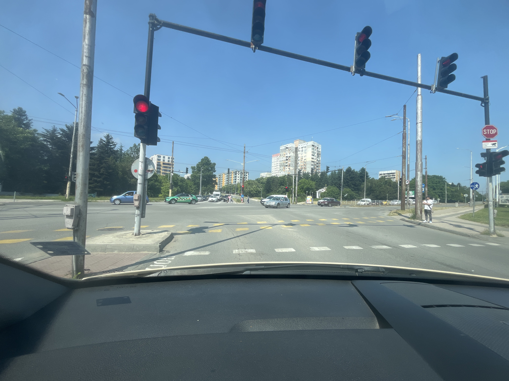 The image displays an urban intersection in Varna, Bulgaria, on a bright, sunny day with a clear blue sky. The perspective is from inside a car, with the vehicle's dashboard visible in the foreground.
The intersection features multiple traffic lights mounted on overhead gantries and poles. The primary traffic light directly above the viewer's lane and several others show a red signal, indicating that traffic is currently stopped.
Several vehicles are visible on the road. A blue car is partially visible to the left, and a green taxi is further ahead. A silver car is stopped in one of the lanes, and a darker car is visible further down the road to the right. Road markings include yellow crosswalk stripes, white dashed lines for lanes, and yellow box junction markings at the center of the intersection.
On the right side of the intersection, a pedestrian, dressed in a white t-shirt and dark pants, stands on the sidewalk, holding a white bag, apparently waiting to cross or proceed.
In the background, several multi-story apartment buildings rise above a dense line of green trees, suggesting a populated, tree-lined urban area. Numerous utility poles and overhead wires crisscross the sky.
Visible text and signs include a red octagonal "STOP" sign with a small icon resembling a surveillance camera beneath it, possibly indicating CCTV monitoring. A sticker with the word "reklama" (Bulgarian for "advertisement") is discernible on a pole. The green taxi also has "TAXI" text, though not fully clear. The scene depicts a typical busy daytime traffic stop at an urban junction.
