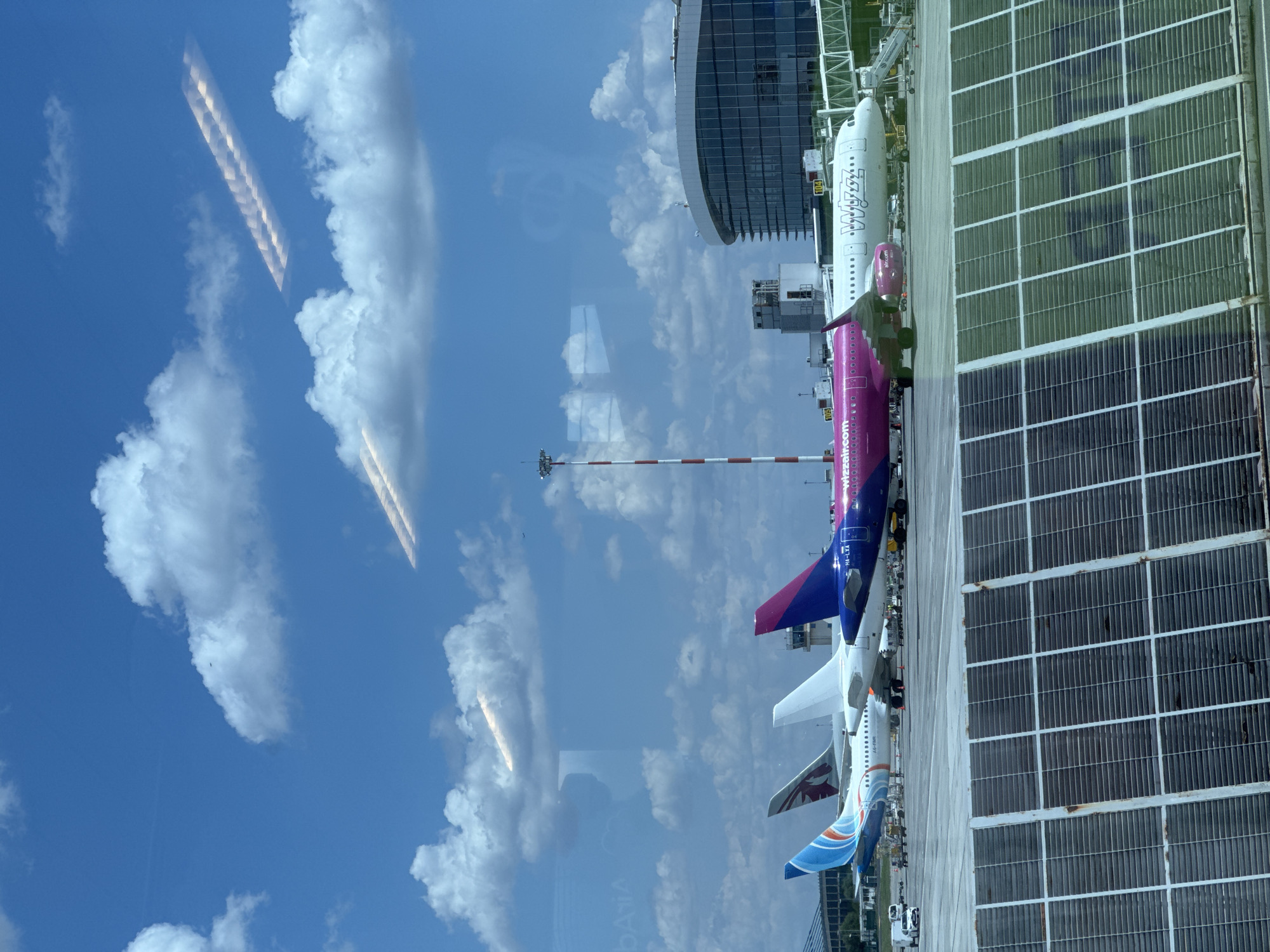 The image depicts an airport tarmac in Otopeni, Romania, under a partly cloudy sky. In the foreground, a section of a solar panel installation is visible, with a grid-like structure and a metallic sheen. Behind this, several commercial airplanes are parked at gates. From left to right, a plane with blue and white livery, possibly a Qatar Airways aircraft, is partially visible. Next to it is a white aircraft, and then a brightly colored pink Wizz Air plane. To the right of the pink plane is a white Wizz Air aircraft. In the background, a modern airport terminal building with large glass windows and a curved roof dominates the scene. A red and white striped control tower stands between the planes and the terminal. The environment suggests a typical operational airport day.