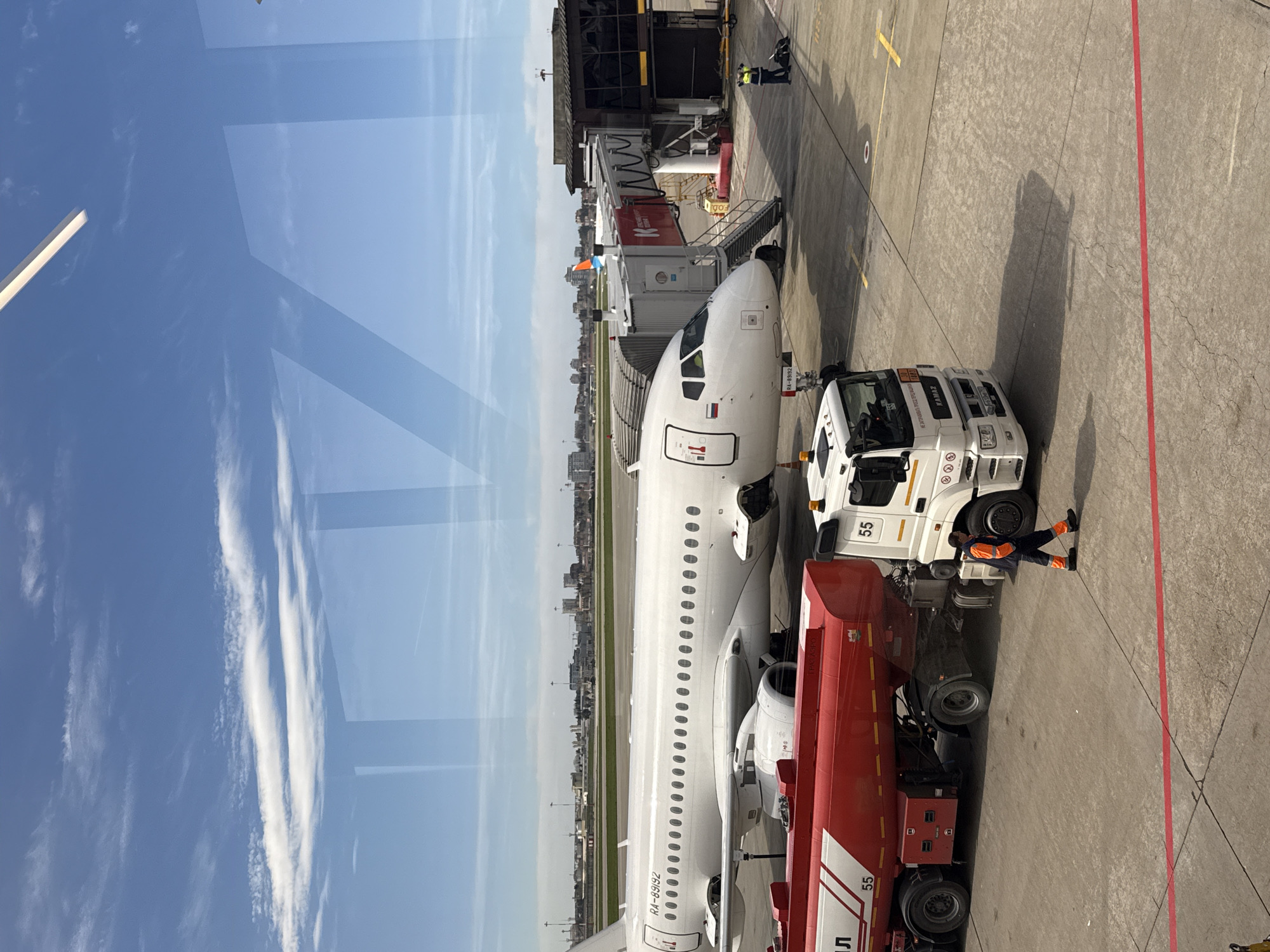 The image depicts an aircraft being refueled at an airport. The scene is from an elevated perspective, likely through a window. A white passenger jet is parked at an airport gate with a jet bridge connected. A red and white fuel truck is positioned near the aircraft, and a worker is present in an orange jumpsuit near the truck. The aircraft's registration number, RA-89192, is visible on the tail. In the background, there's an urban area, and the sky is blue with clouds. Based on the aircraft livery and the presence of a flag, it is possible that this is located in Sochi, Russia.