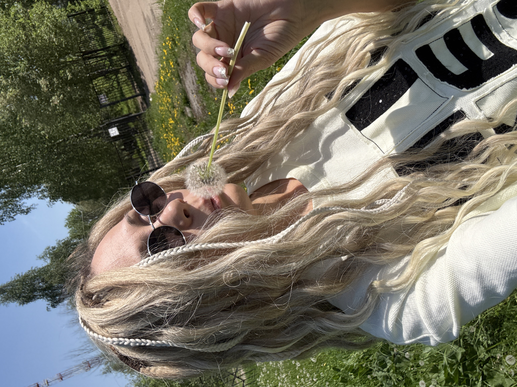 The photograph captures a person lying on the grass in an outdoor setting, with patches of green and dandelions visible. The individual has long, blonde, braided hair and wears round sunglasses. They are holding a dandelion in front of their face. The person is wearing a white garment with black patches or embellishments. The location could potentially be near Шебунино, Russia, given the mention of the city.