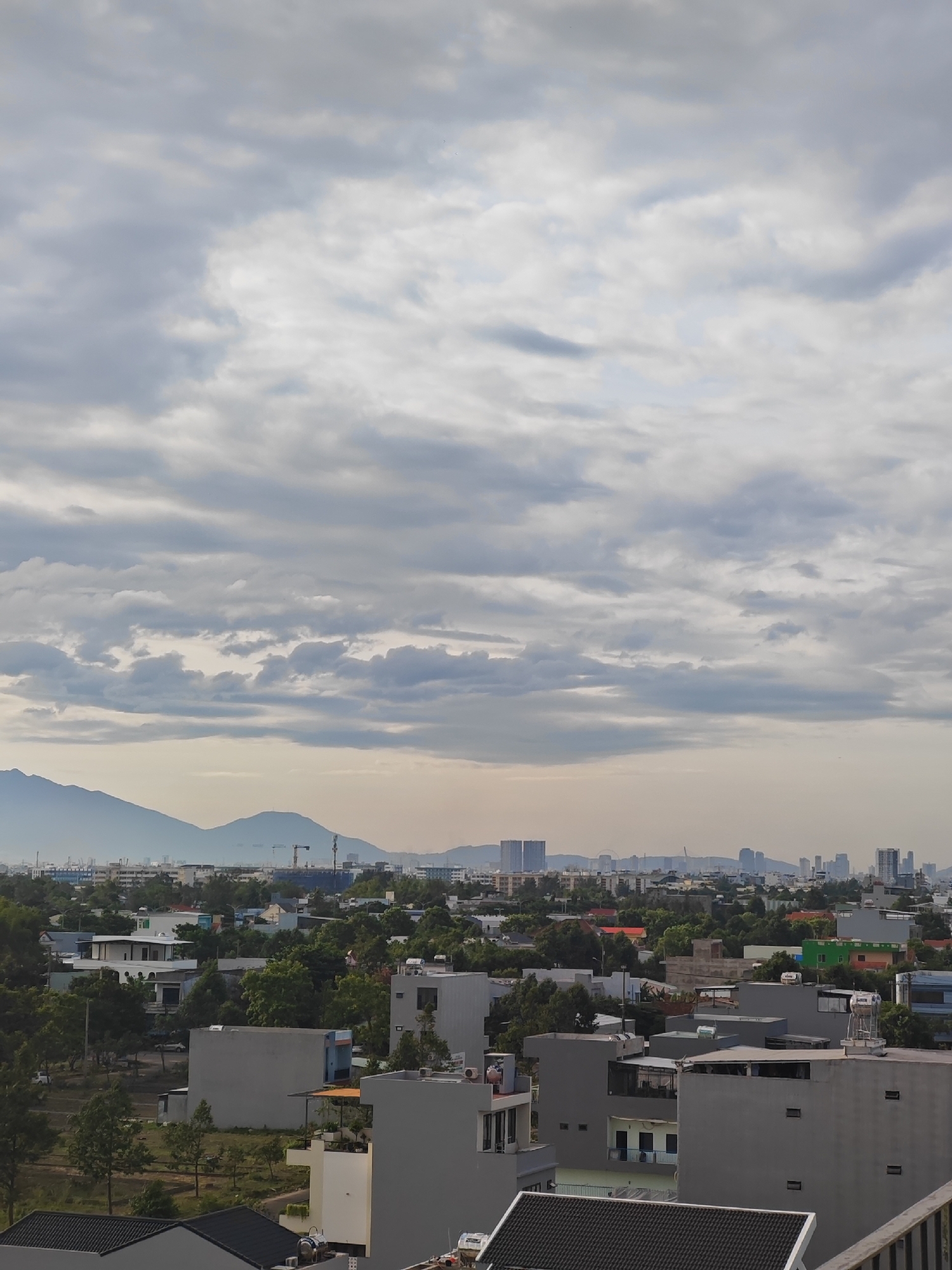 The image depicts an aerial view of a densely populated urban area, likely within the city of Đà Nẵng, Vietnam, characterized by a mixture of residential buildings, scattered trees, and distant mountains under a cloudy sky. The foreground features several multi-story residential buildings, predominantly in shades of gray, white, and light beige, with flat roofs and visible air conditioning units. Some buildings display exposed piping and water tanks. The middle ground is a sprawling expanse of more residential structures, interspersed with lush green trees and some smaller commercial buildings, identifiable by their colorful roofs, such as a bright green one. Cranes indicate ongoing construction in various parts of the cityscape. In the background, a hazy skyline of taller buildings and what appear to be skyscrapers is visible, with rolling green mountains forming the horizon. The sky is overcast with a layer of grey and white clouds, suggesting either early morning or late afternoon conditions, with soft, diffused light. No people are visible in the frame.