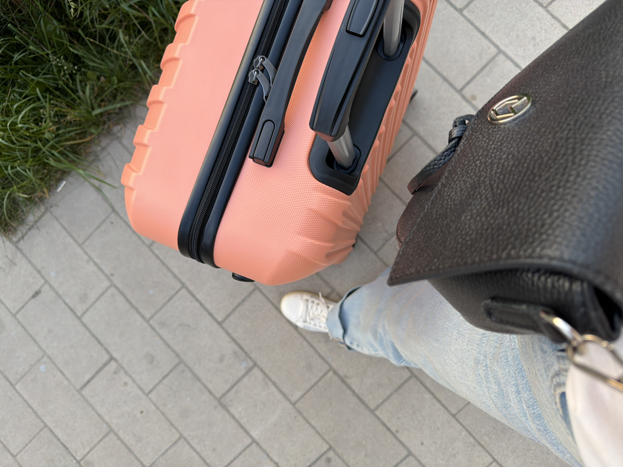 A first-person perspective shot shows a person's legs and feet on a paved sidewalk. They are wearing light blue denim jeans and white sneakers. To the right of the person's legs is a coral-colored suitcase with black trim and a black handle. The suitcase has a textured surface and appears to be upright. In front of the suitcase and to the left of the person's legs is a dark brown leather handbag. The handbag has a textured surface and a metallic clasp with an embossed logo. The background consists of the paved sidewalk and a strip of green grass on the right side of the frame. There are no visible landmarks or specific architectural features to indicate a location within Moscow, Russia.