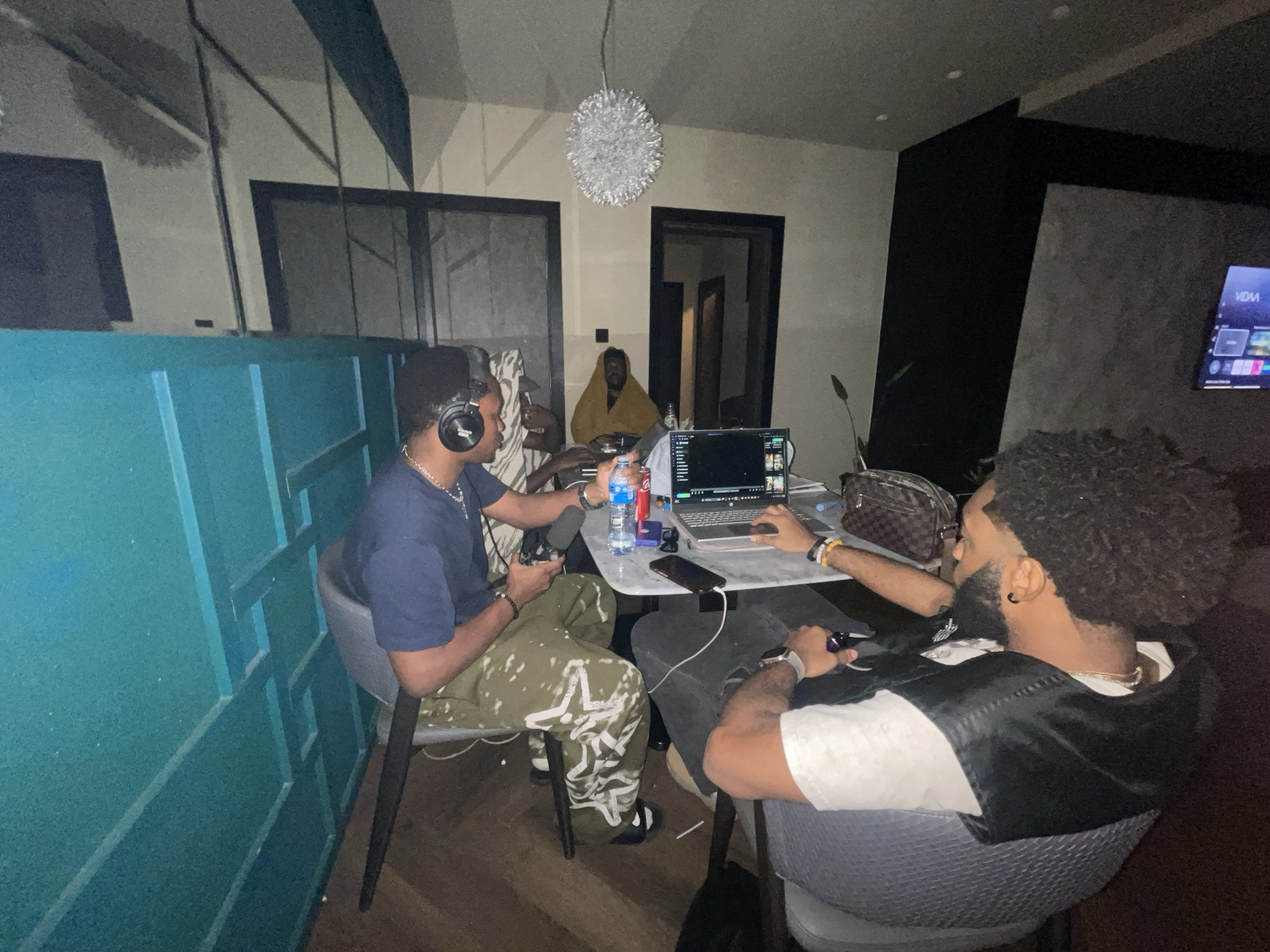 A group of four men are gathered in a room, possibly in Abuja, Nigeria. The room's décor features a teal-colored wall with paneling, a dark accent wall with a television, and a modern chandelier. The men are seated around a light-colored marble-topped table. One man, wearing headphones, a navy-blue shirt, and patterned green pants, holds a video game controller. Another man, with an afro hairstyle and wearing a white t-shirt with a black vest, also holds a video game controller. A laptop sits on the table between them, along with a water bottle, soda can, and other personal items. A third man is partially visible in the background, wearing a gold/brown hooded shawl. The room has multiple doorways, and the overall lighting is dim.