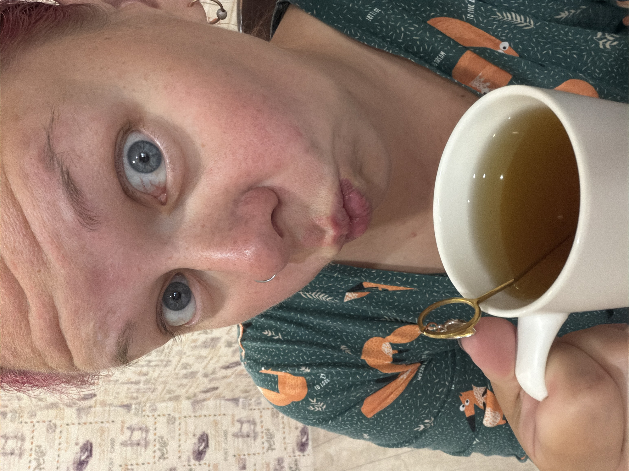 A woman with pink-tinted hair is holding a white mug of tea, the mug is to the right of the frame. She is looking at the camera with wide blue eyes and her lips are pursed. She is wearing a green shirt with an orange fox pattern. The background is a wall covered in a patterned wallpaper. The image is a close-up, so the location in the city of Chaikovsky, Russia is not discernible.