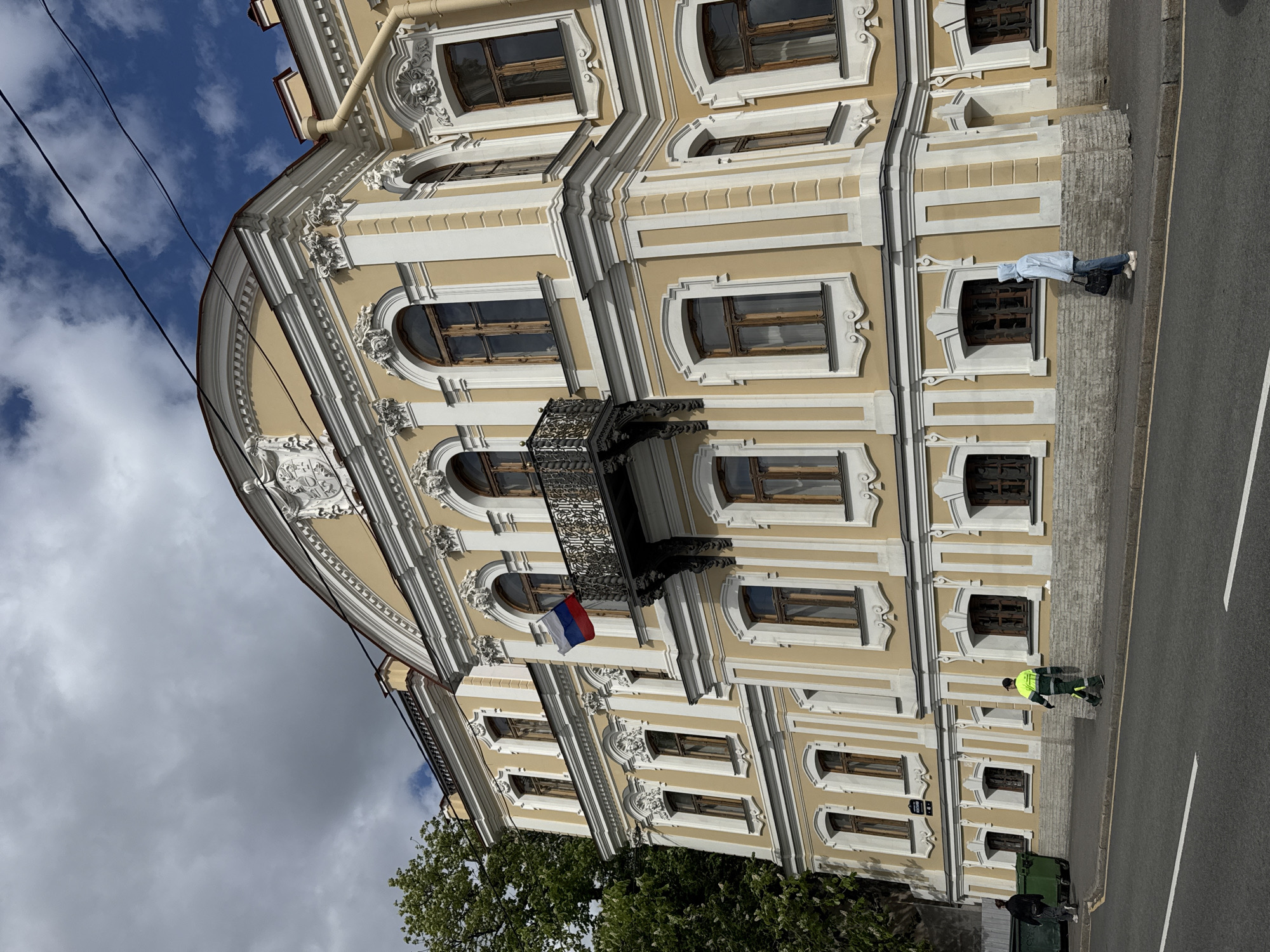 The image shows the facade of a building in Saint Petersburg, Russia. The building is primarily light yellow with white architectural details around windows and along the edges. It has multiple arched windows on each level. A wrought-iron balcony is visible on the upper level. The Russian flag is displayed from the balcony. On the sidewalk in front of the building, there are two individuals: one person wearing a light blue shirt and jeans, and another person dressed in a reflective neon green and black work uniform. A street can be seen on the right side of the image. Trees and cloudy skies can be seen in the background.