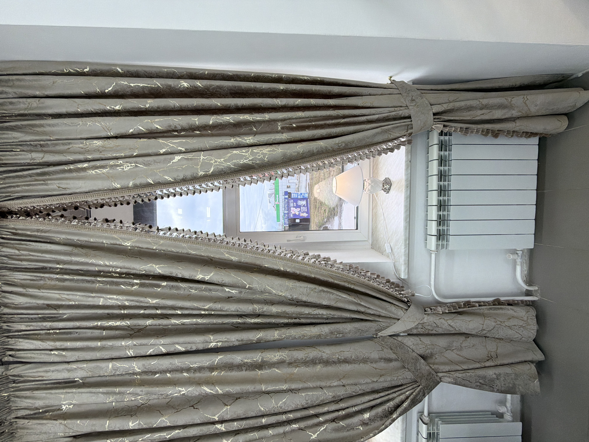This image captures an interior view focusing on a window dressed with elegant curtains. The curtains are made of a rich, taupe or grayish-brown fabric adorned with a distinctive gold, marble-like, or veined pattern. They are drawn back to both sides, forming deep pleats and held in place by matching fabric tie-backs. A decorative fringe or tassel trim in a complementary color runs along the bottom edge of the curtains where they frame the window.
Through the white-framed window, a bright outdoor scene is visible, indicating it is daytime. The exterior view includes distant buildings, what appears to be a street or parking lot, and some sparse vegetation. A prominent blue sign outside displays Cyrillic text, clearly showing "ЦВЕТЫ И БУКЕТЫ" (Flowers and Bouquets), with additional text "ВМЕСТЕ" (Together) visible below it, suggesting a business in the city of Votkinsk, Russia.
On the white windowsill, a small, decorative table lamp with a white shade and an ornate base is positioned. Below the right side of the windowsill, a modern white radiator with vertical panels is installed against a plain wall. The scene is static, showcasing the detailed window treatment and a glimpse of the exterior. No people are present in the image.