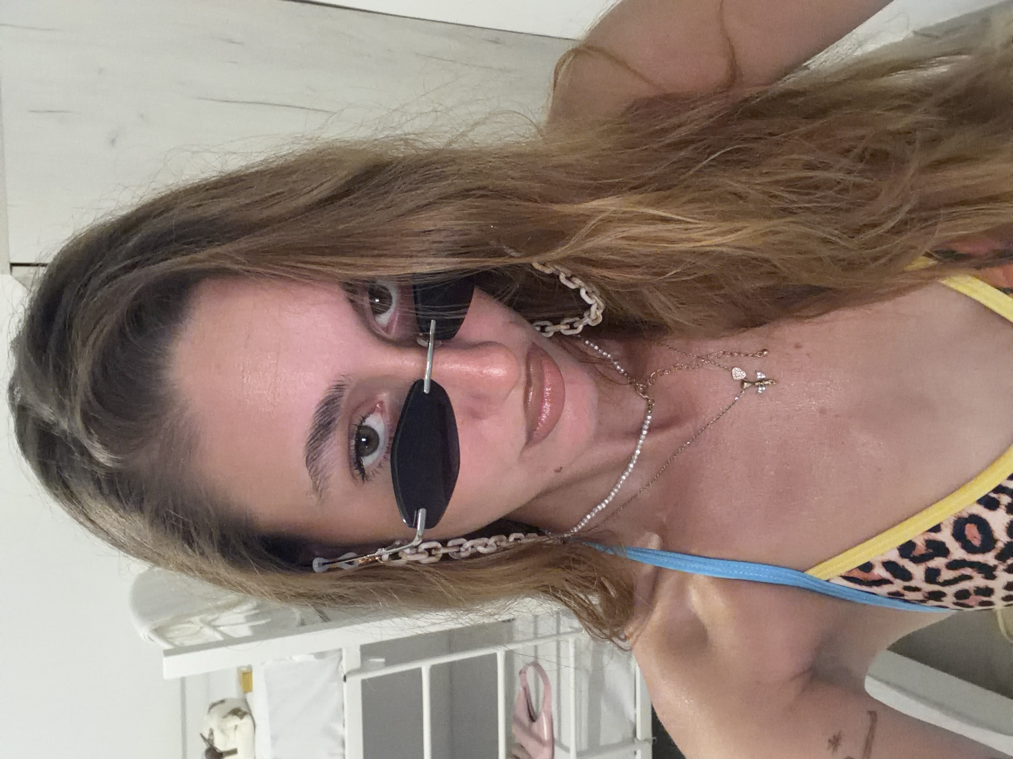 A woman with long, wavy blonde hair and fair skin is pictured from the chest up. She is wearing black, oval-shaped sunglasses with a chain attachment, a multi-layered necklace, and a leopard-print bikini top with blue and yellow trim. The background appears to be an indoor setting, possibly a bedroom or dorm room, with a white bunk bed structure visible on the left and a white wooden door on the right. The image is a selfie taken from a low angle. There is no indication of the location being Stavros, Greece.