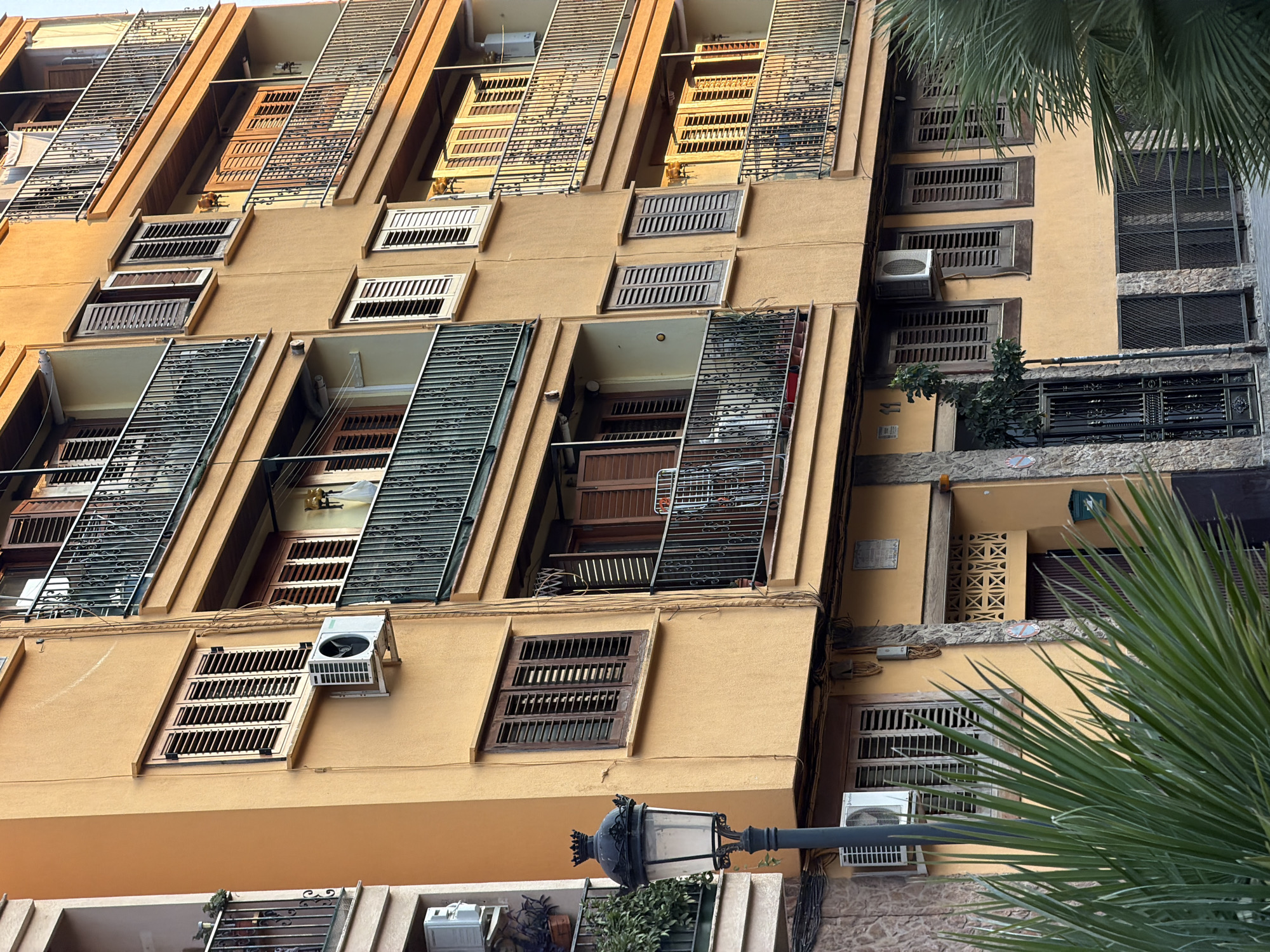 The image shows a multi-story apartment building in Benimàmet-beniferri, Spain. The building has an orange-yellow facade with several balconies featuring black metal railings. The windows are fitted with brown wooden shutters, some of which are open, revealing interior details. Air conditioning units are visible mounted on the exterior walls below some of the windows. A black vintage-style street lamp stands in the foreground to the left, partially obscured by lush green palm fronds. The scene is illuminated by warm sunlight, suggesting it was taken during the late afternoon or early morning. The building appears to be in a residential area.