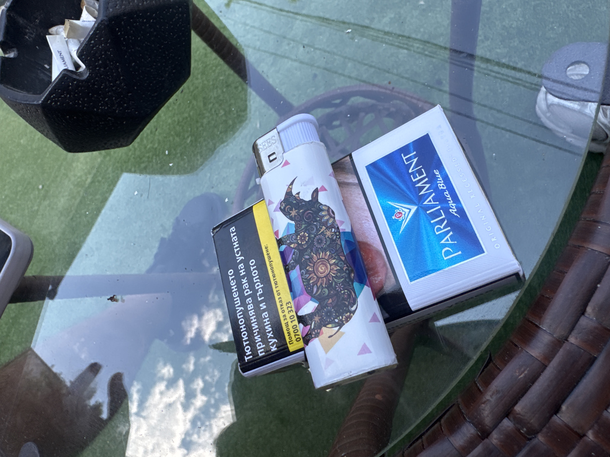 An overhead shot captures smoking paraphernalia arranged on a glass-top table in what appears to be an outdoor or semi-outdoor setting in Varna, Bulgaria. The sun is shining, and reflections of white clouds against a blue sky are visible on the table surface.
The main subjects include a textured black ashtray containing several discarded cigarette butts, some with the brand "LAMENT" visible. Next to it, a white disposable lighter featuring a distinctive colorful rhinoceros design with intricate patterns is positioned. Part of the lighter's branding, "SEES," is legible. Adjacent to the lighter is a pack of "PARLIAMENT Aqua Blue" cigarettes, with the text "ORIGINAL RECESSED FILTER" partially visible. Another, mostly obscured cigarette pack is beneath the lighter.
The visible side of the cigarette pack displays health warnings in Bulgarian: "Тютюнопушенето причинява рак на устната кухина и гърлото" (Smoking causes oral and throat cancer) and "Помощ за отказ от тютюнопушене: 0700 10 323" (Help to quit smoking: 0700 10 323).
Beneath the transparent glass table, a green, possibly artificial grass, surface is visible, along with dark brown wicker-patterned furniture, likely a chair or table base. The arrangement suggests a casual break or leisure time.