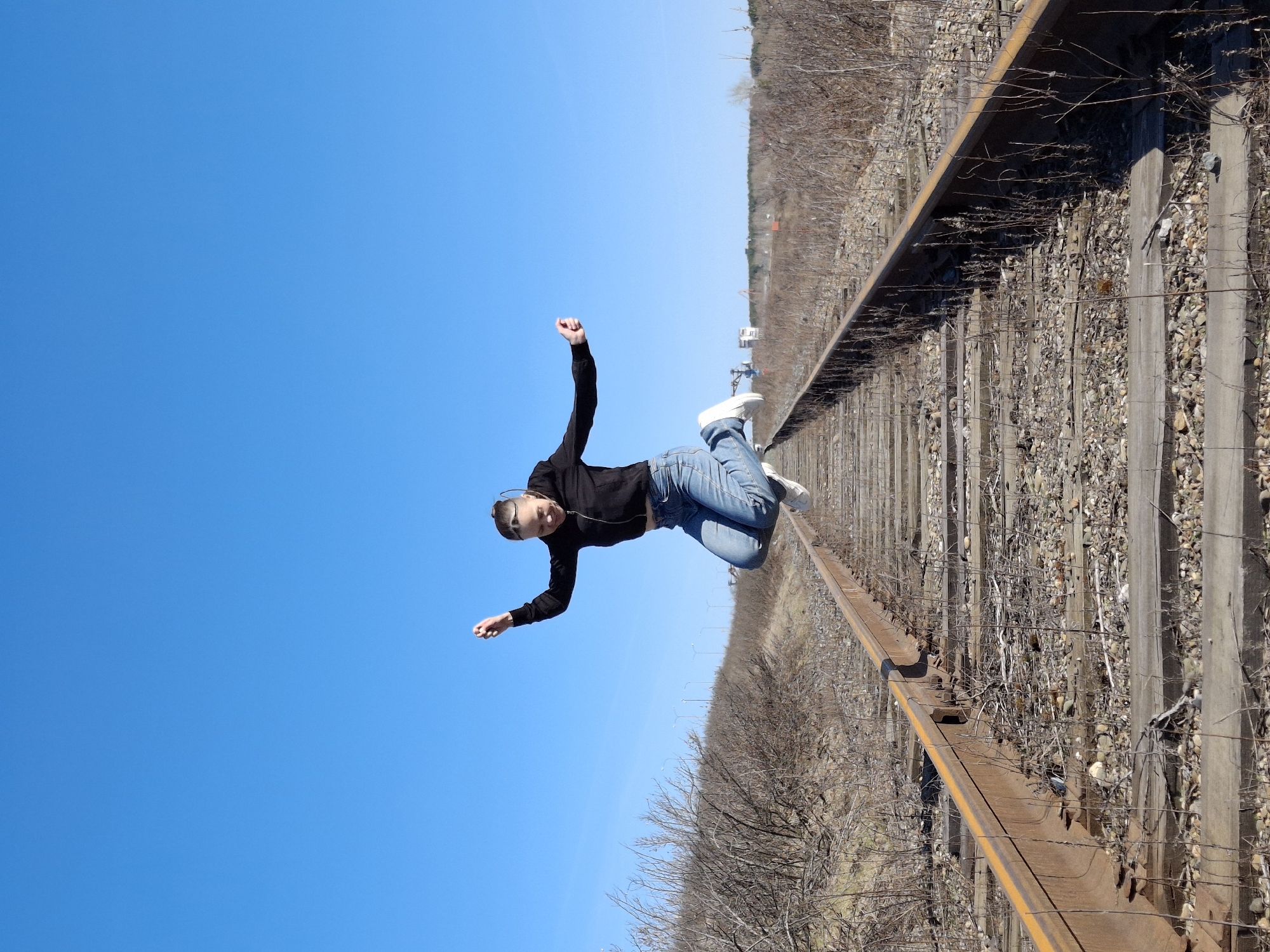 The image displays a young person, likely female, suspended in mid-air against a clear blue sky, with railroad tracks and a barren landscape positioned perpendicular to their body. The overall photograph appears to be rotated 90 degrees counter-clockwise.
The subject is dressed in a black long-sleeved top, blue jeans, and white sneakers. Their hair is pulled back, and sunglasses are perched on top of their head. They are captured mid-jump or mid-fall, with arms bent and hands slightly open, and legs bent at the knees. Their expression is concentrated, with their tongue slightly out, suggesting a playful or artistic pose rather than an accidental fall.
The background on the left and upper side of the rotated image is a vast, cloudless, and bright blue sky. To the right and bottom, a single set of rusty railroad tracks extends diagonally into the distance. The tracks are laid on gravel and a mix of weathered wooden and concrete sleepers. The area immediately surrounding the tracks features sparse, dry vegetation, primarily bare bushes and trees, characteristic of an early spring or late autumn landscape in Chaikovsky, Russia. Bright, direct sunlight indicates a midday setting, casting visible shadows. There is no visible text present in the image.