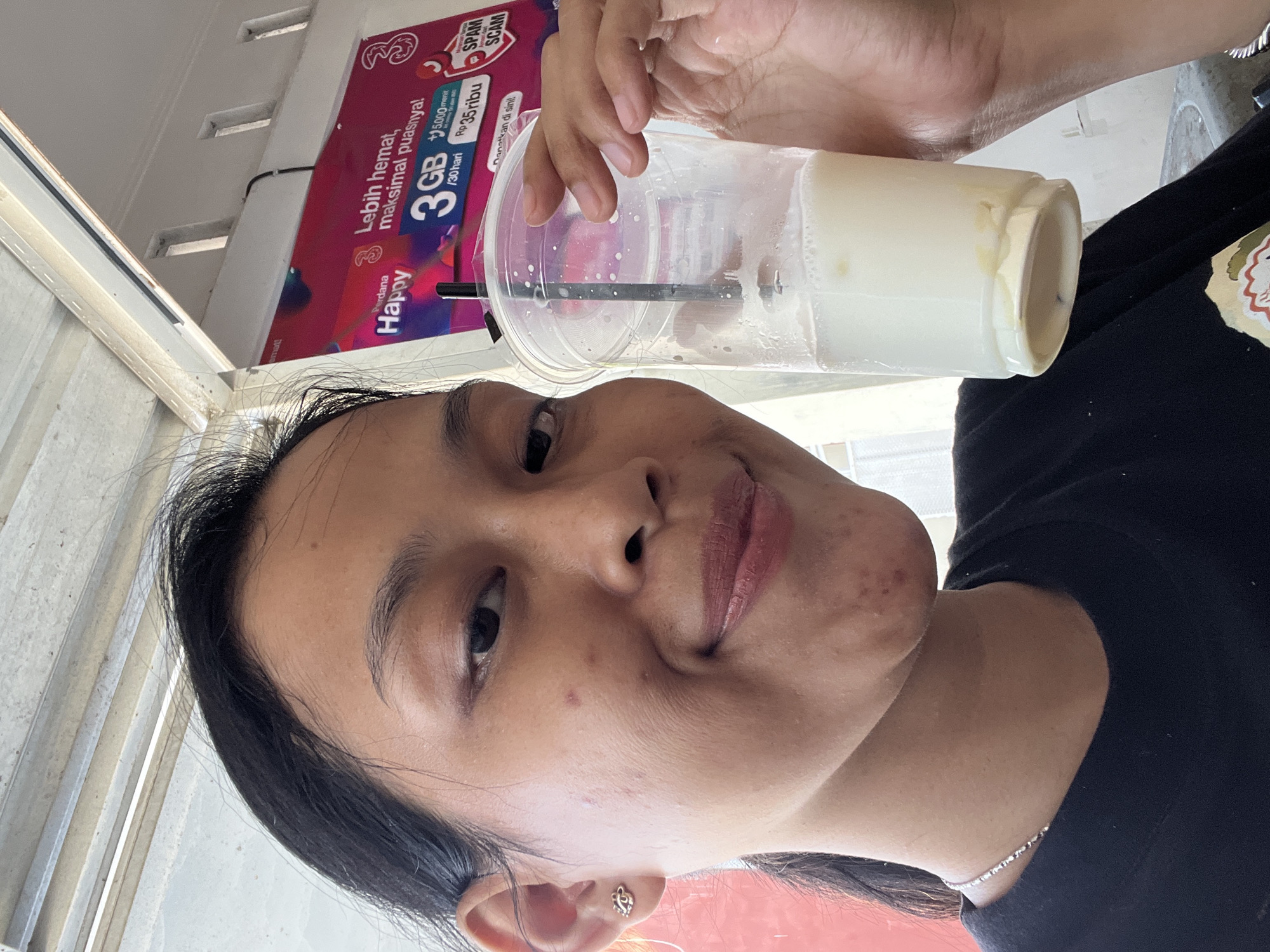 A young woman with dark hair pulled back is holding a clear plastic cup with a white beverage and a black straw. She is wearing a black t-shirt and a silver necklace. Her head is tilted slightly, and she is looking at the camera with a smile. She has on light-colored makeup, including lipstick. The background shows a white ceiling with metal supports and a pink advertisement banner with text in Indonesian. The overall lighting suggests an outdoor or semi-outdoor setting.