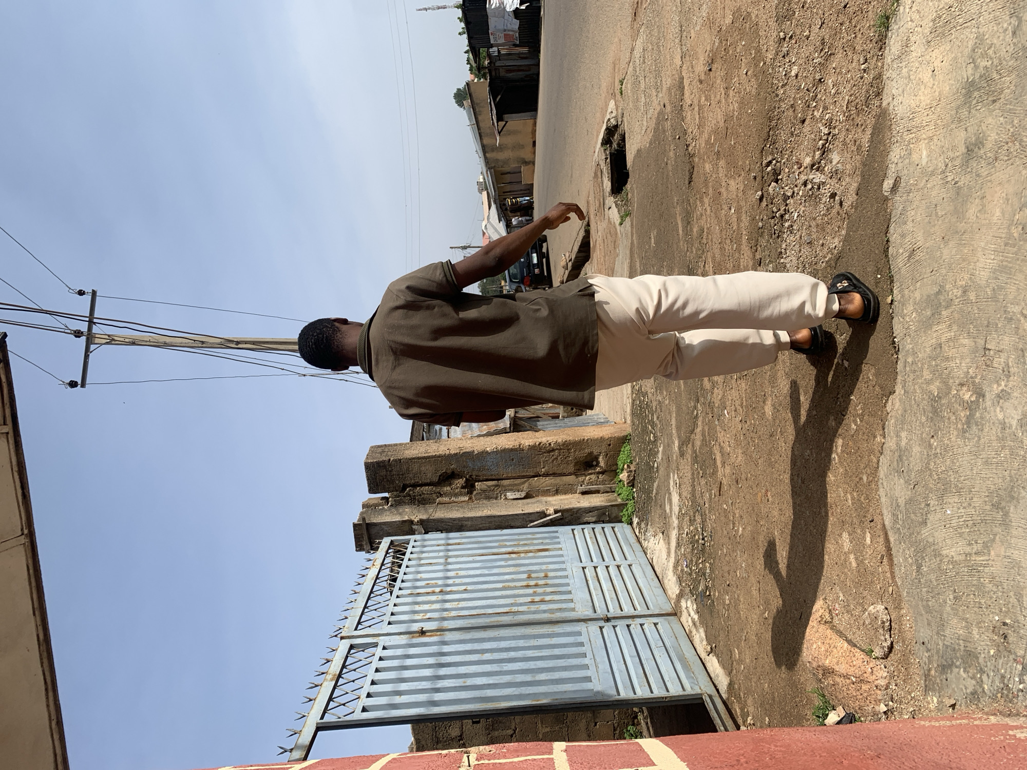The image depicts a man walking away from the camera on a dirt and concrete pathway next to a street in Gura Top, Nigeria. The man is wearing a brown short-sleeved polo shirt and light-colored pants. He is wearing dark sandals. To his left is a blue metal gate with vertical slats and pointed spikes along the top. Behind the gate are brick structures. A utility pole with wires runs diagonally across the sky. The street is paved and has buildings and structures along its right side, some with corrugated metal roofs. A dark-colored car is parked on the left side of the street. The ground is a mix of dirt, gravel, and concrete. The sky is clear and blue.