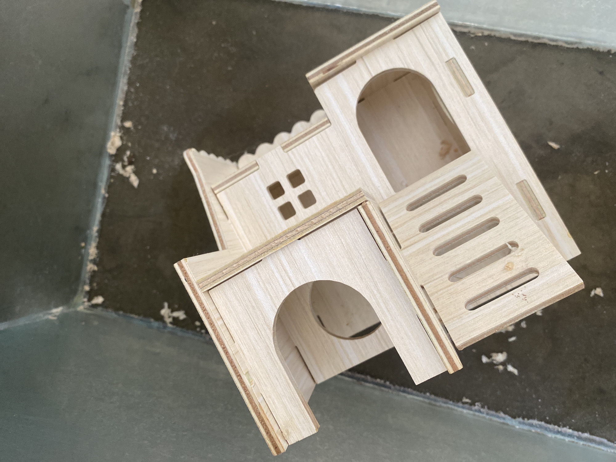 An unfinished wooden model of a multi-level house, possibly for a pet or a craft project, is placed on a dark, textured surface. The model features several openings, including a large circular entrance and a smaller arched doorway, as well as small square window cutouts and a slatted wall. The wood appears to be plywood or a similar material, with visible grain and layering. The surrounding environment appears to be a floor with some debris and a wall in the background. The location is identified as Davao City, Philippines.