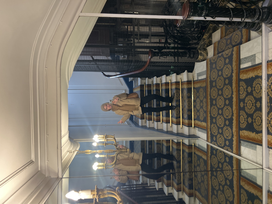 A person wearing a beige coat, scarf, and hat is standing on a carpeted staircase. They are holding a smartphone and pointing. Behind them is an elevator shaft. The staircase is flanked by walls. The image is reflected in a mirrored surface along the bottom edge, creating a duplicate scene with light fixtures visible in the reflection.