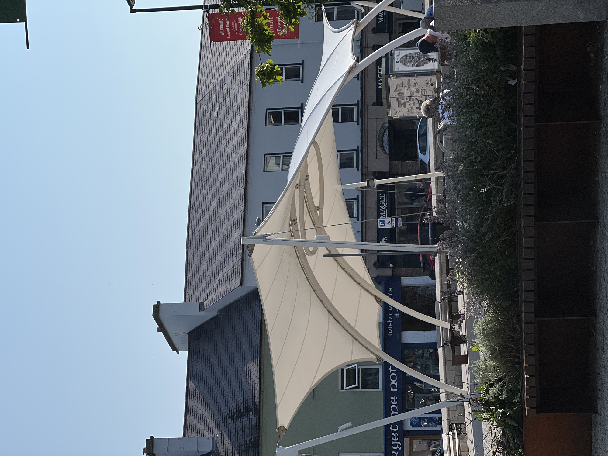 This is a photograph taken on a clear, sunny day in Donegal, Ireland, likely in the town square. In the foreground, there are several benches and a planter filled with lush, green and purple foliage. Behind the greenery, a modern, white, tensile fabric canopy structure is supported by white poles, casting shadows on the ground.
Beyond the canopy, a street-level view of shops and buildings is visible. One shop has a sign reading "Forget Me Not." Another store has signage for "Irish Crafts." Further along, there are storefronts with signs like "Magee 1866" and "Donegal Tweed." The buildings appear to be two or three stories tall, with a mix of windows and architectural styles, some with grey slate roofs and chimneys.
In the middle ground, several people are seated on benches or standing near the shops. One person is reading a book, and another appears to be wearing a blue shirt and a cap. A car is parked in front of one of the shops.
In the background, the sky is a clear, bright blue. A red banner is hanging from a lamppost, displaying text in multiple languages including "WILKOMME" and other greetings. The overall impression is a lively, public space in a coastal Irish town on a pleasant day.