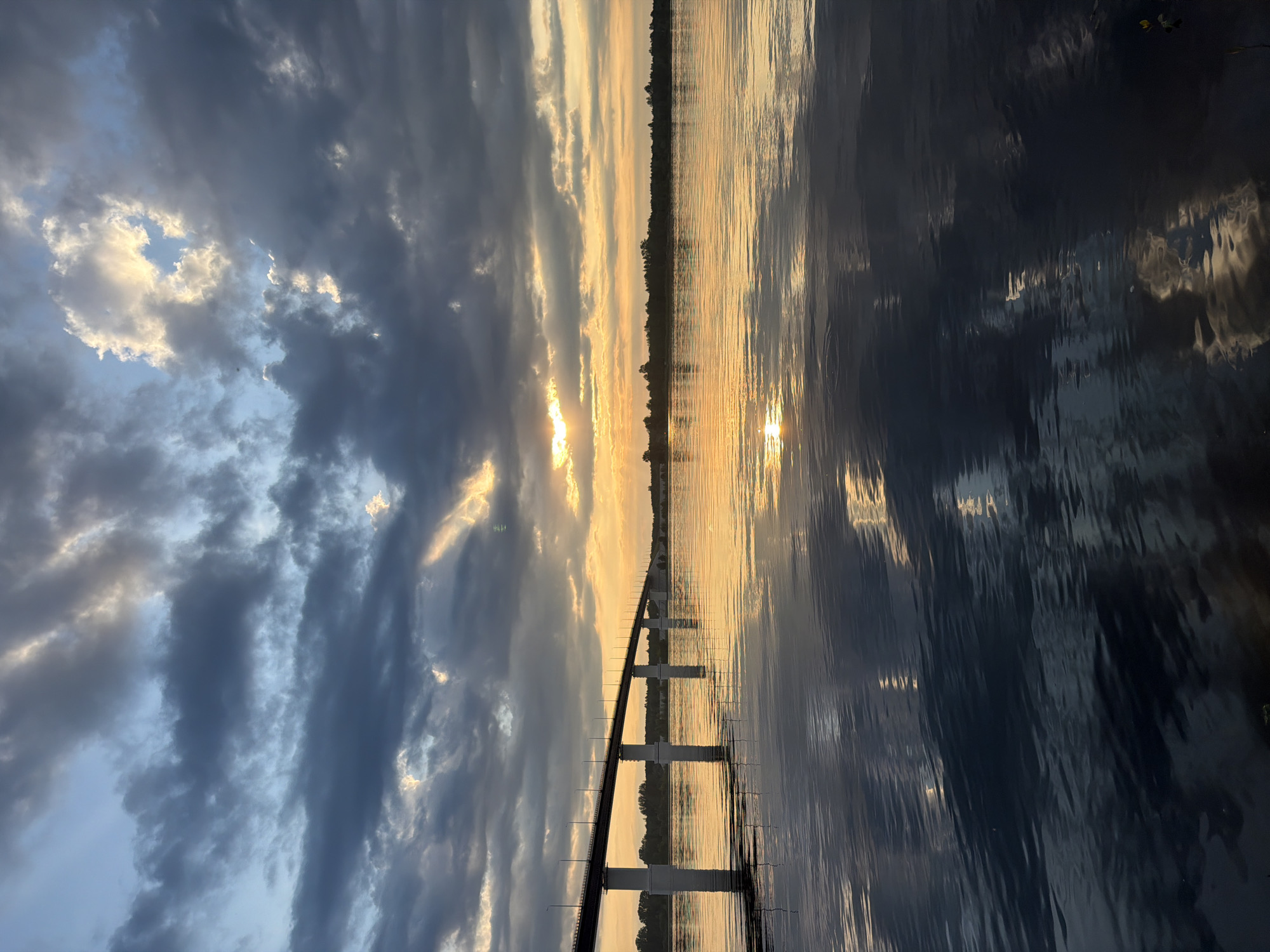 The image depicts a scenic view of a wide river with a bridge crossing it at sunset. The sky is filled with dramatic clouds, some dark and stormy, others illuminated by the setting sun, casting a warm orange glow across the water. The bridge, supported by several piers, stretches across the river, with its reflection visible on the water's surface. The far bank of the river is lined with trees, creating a dark silhouette against the vibrant sky. The water itself is calm, with ripples reflecting the colors of the sky and the bridge. The scene is tranquil and picturesque, capturing the beauty of a sunset over a river in Kambarka, Russia.