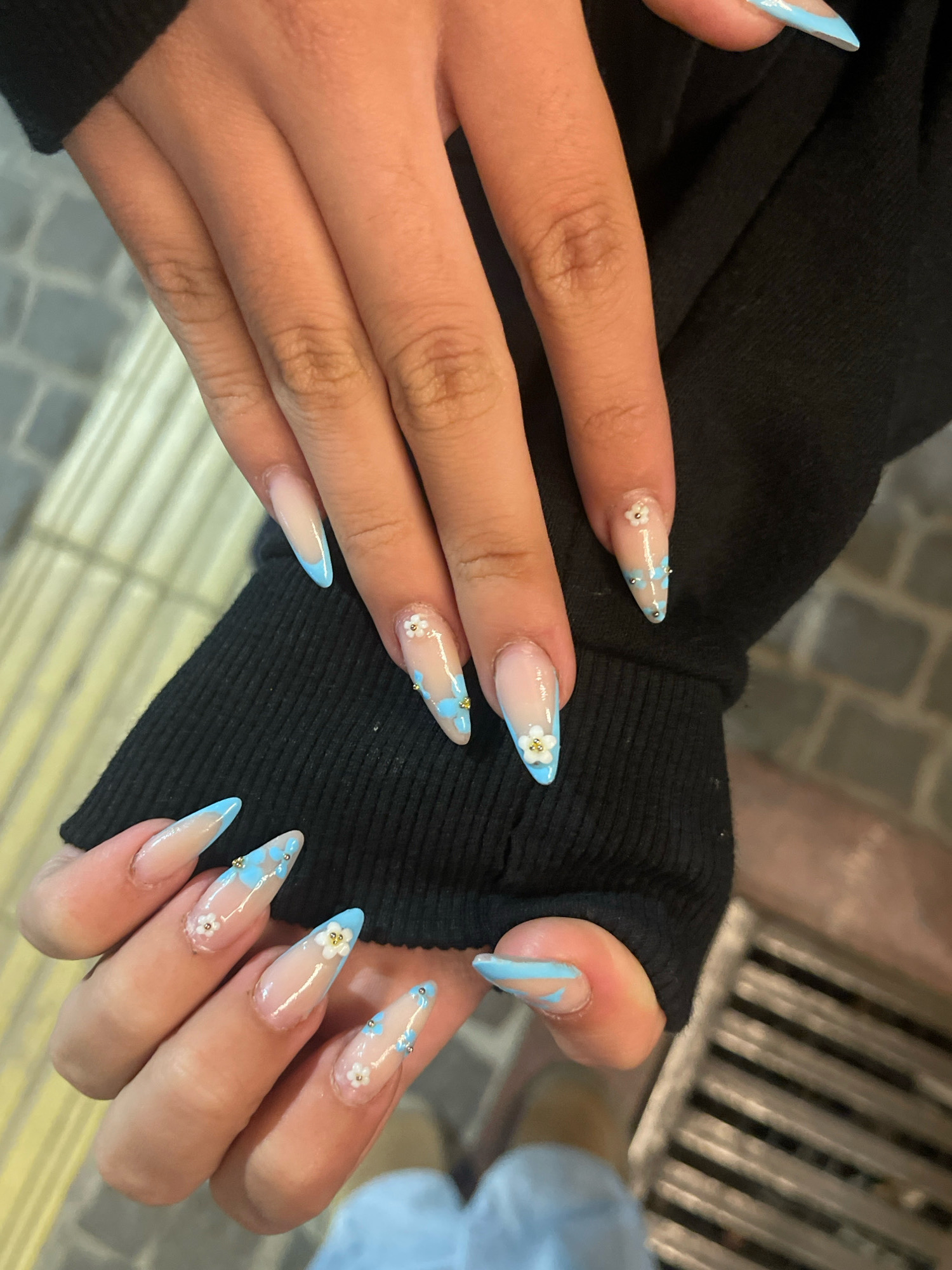 In Prizren, Kosovo, a close-up shot captures a person's hands adorned with long, almond-shaped acrylic nails. The nails feature a gradient design, transitioning from a sheer, natural base to a light blue tip. Scattered across several nails are delicate white floral embellishments with small gold accents, along with tiny blue flower-shaped decals and small gold bead details. The person is wearing a black long-sleeved garment, with the sleeves of the garment visible. The background suggests an outdoor urban setting, with cobblestone pavement and metal grates visible, typical of Prizren's historic city center.
