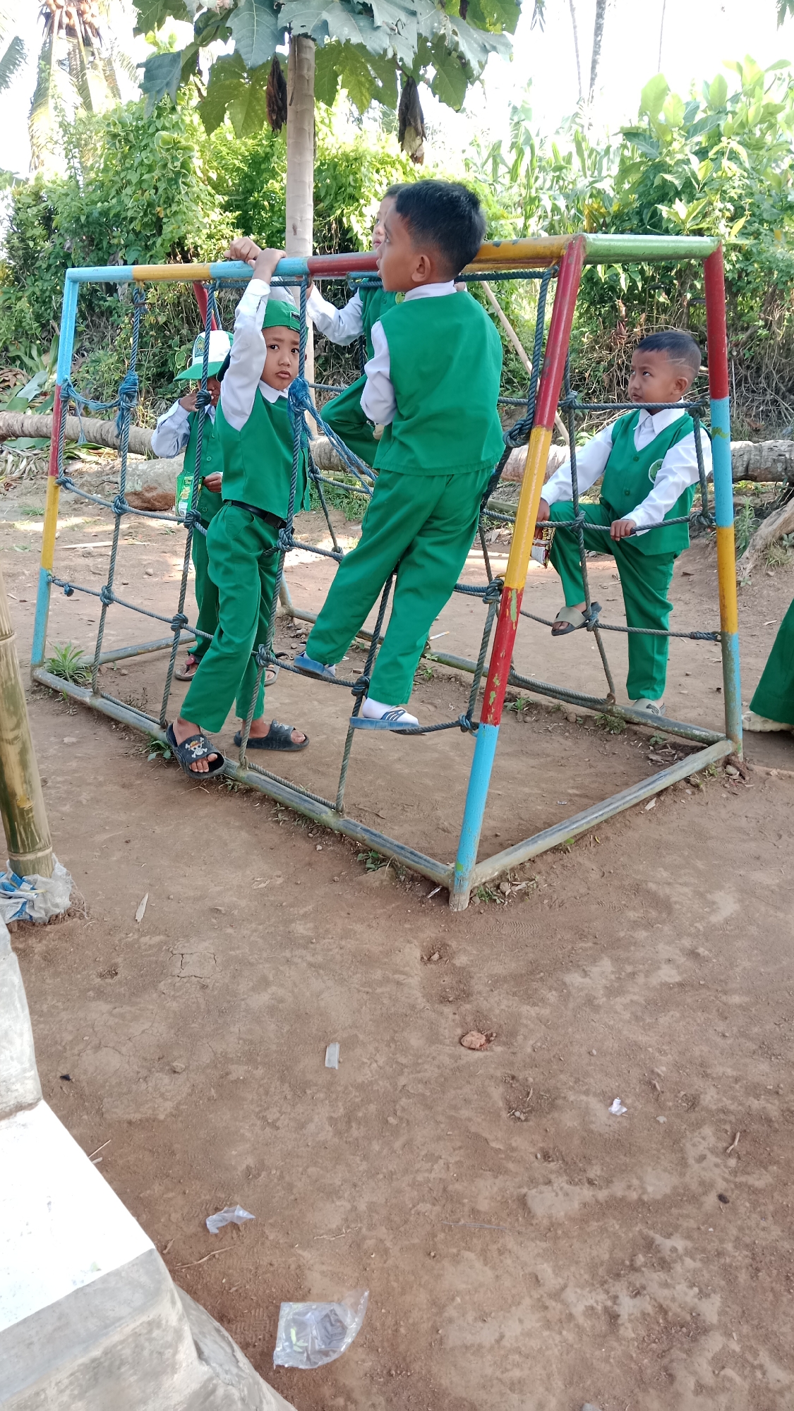 Four young children, dressed in matching green vests and pants over white shirts, are playing on a colorful climbing structure in an outdoor setting. The structure is made of metal poles painted red, yellow, and blue, with a rope net for climbing. The ground beneath is dirt. In the background, there is lush green vegetation, including trees and foliage, suggesting a rural or park-like environment. The scene appears to be in the city of Karangharjo, Indonesia, based on the provided context. The children are actively engaged, with one child hanging from the structure, another standing on a rope, and two others positioned at different levels. The lighting suggests it is daytime with sunlight.
