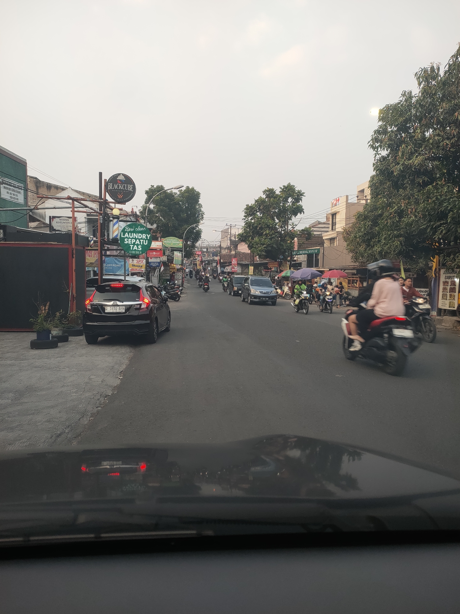 The image depicts a street scene in Cimahi, Indonesia, likely during daylight hours given the ambient light, though the sky is overcast. The perspective is from inside a vehicle looking forward. The street is paved and appears to be a busy thoroughfare with moderate traffic.
Several vehicles are visible: a black Honda hatchback with license plate D 1610 AHN is prominent in the foreground on the left side of the road. Further down the road, other cars, including a dark minivan, are present. A significant number of motorcycles are also navigating the street, some carrying one or two riders. A few individuals are visible on the motorcycles, wearing helmets and casual clothing. One person in the foreground on a red motorcycle is wearing a light pink shirt.
Along the sides of the street, there are various buildings and businesses. Signs are visible, including "BLACKCUBE BARBERSHOP", "LAUNDRY SEPATU TAS", and other commercial advertisements. Some of these signs are mounted on poles, and one business appears to be a laundry service. Trees and foliage are present, adding greenery to the urban environment. The overall impression is a typical commercial and residential street in a moderately populated Indonesian city. The atmosphere appears to be one of daily activity and movement.