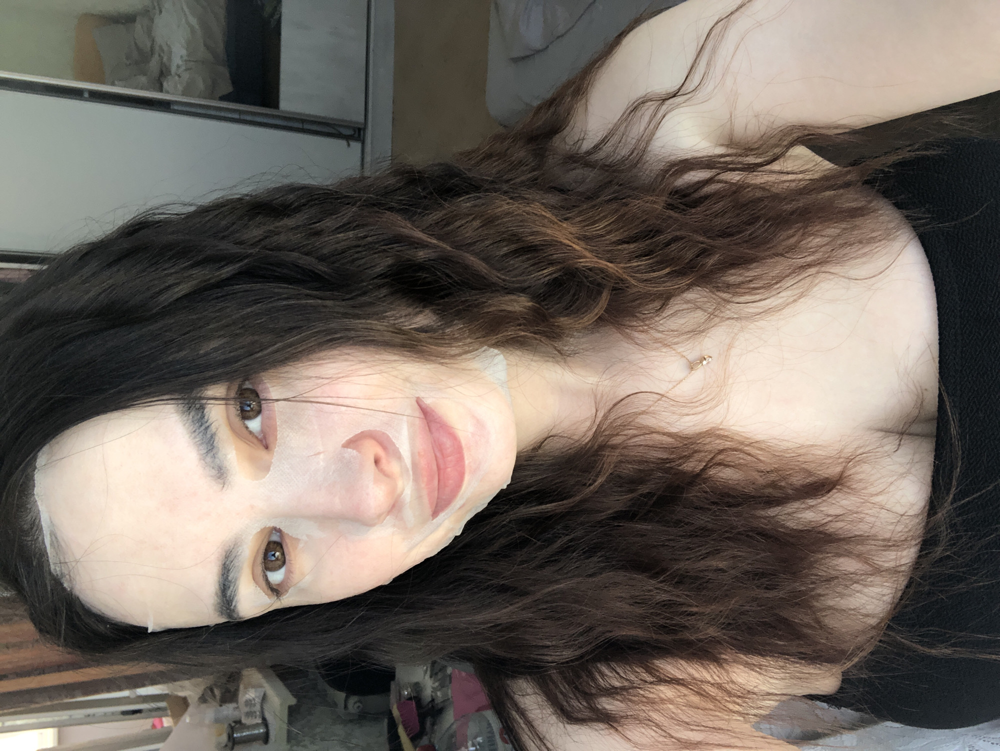 The image depicts a woman with long, dark brown wavy hair wearing a white sheet mask on her face. The mask covers her forehead, nose, and mouth. She is wearing a black top. A small gold necklace is visible around her neck. The background appears to be an indoor space, possibly a bedroom, with a cabinet or wardrobe visible. The location of Noviy, Russia, cannot be confirmed.