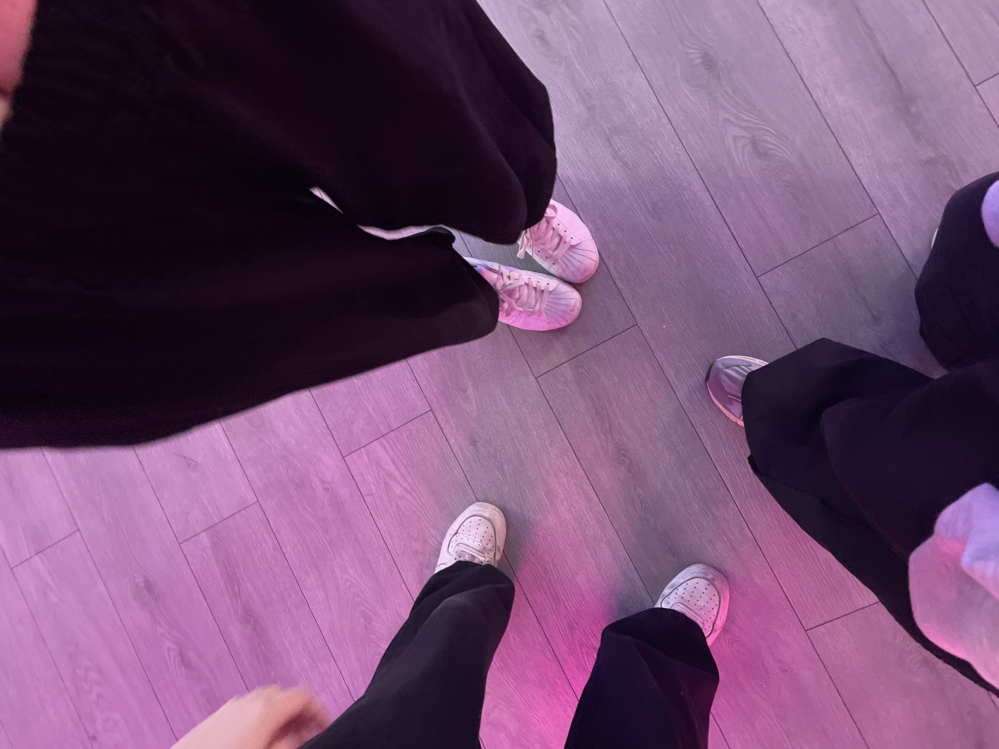 The image, taken in Saint Petersburg, Russia, presents an overhead view of the lower halves of three individuals standing on a light-colored wooden floor. The floor is cast in a prominent pink and purple glow, suggesting artificial lighting, possibly from LEDs, creating a distinct ambiance.
In the upper left quadrant, one person wears a dark, possibly black, flowing garment, such as wide-legged trousers or a skirt. Their feet are clad in light pink or purple sneakers with white soles and laces, appearing to be a classic sportswear style.
At the bottom center, another individual is visible, wearing dark, likely black, trousers and white sneakers. These shoes feature a perforated toe box and a substantial white sole, characteristic of popular sneaker models.
On the right side of the frame, the leg and foot of a third person can be seen. They are also wearing dark, presumably black, trousers and silver or light grey sneakers.
The individuals appear to be stationary, possibly posing for the photograph in an indoor setting like an event space, club, or a room with creative lighting. The intense color wash from the lighting is a key visual element. No discernible text or explicit branding is visible on the clothing or the floor. The lighting suggests an evening or night setting indoors.