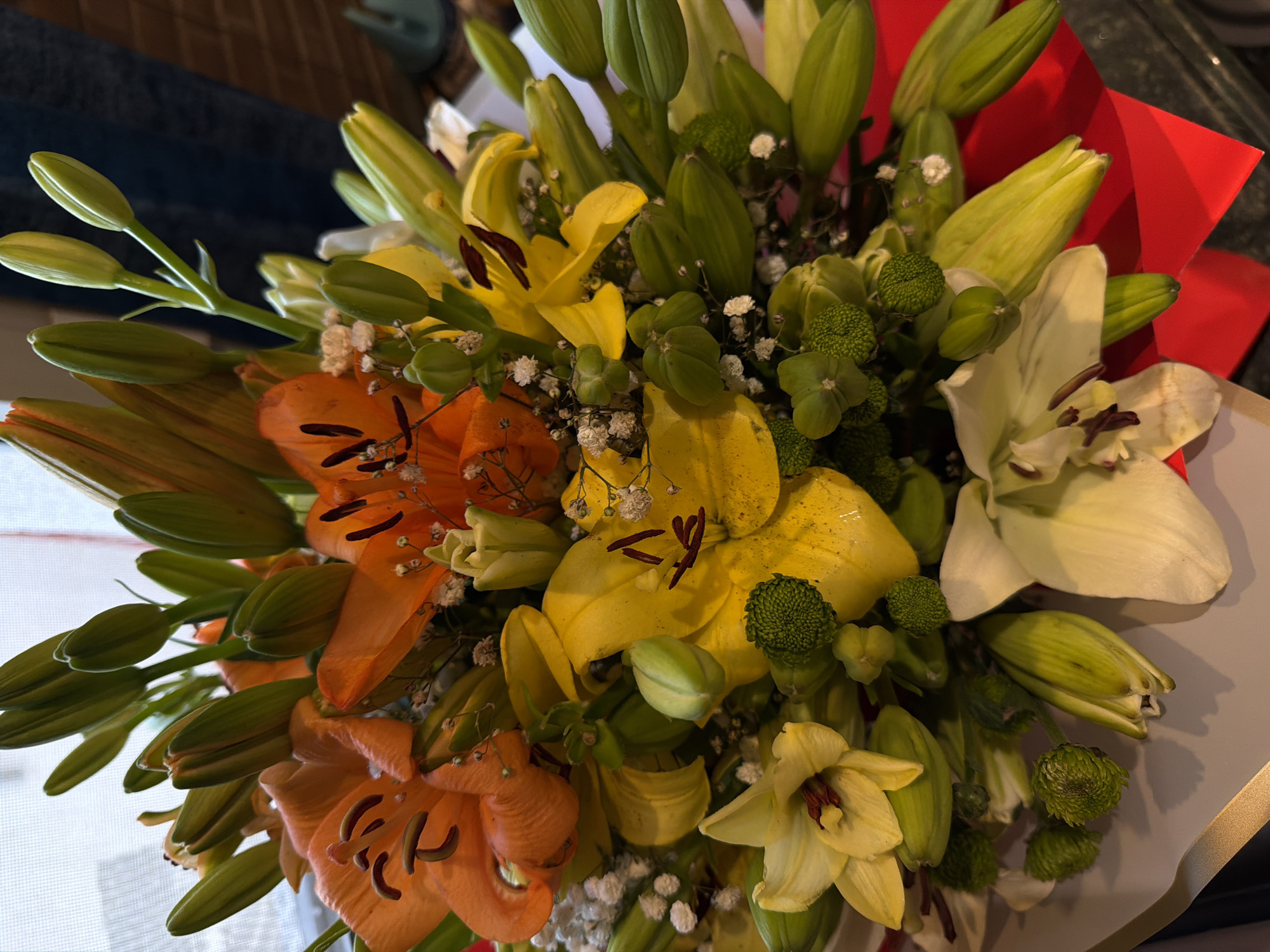 A close-up, overhead shot showcases a vibrant bouquet of lilies, predominantly in orange, yellow, and white hues, interspersed with green lily buds. The flowers are densely packed, with some in full bloom and others still tightly closed. Delicate white baby's breath flowers and small green decorative elements add texture to the arrangement. The bouquet is partially wrapped in red paper and a light-colored paper or ribbon. The background is blurred, suggesting an indoor setting with diffused light, possibly near a window with a textured blind. There is no visual information to suggest the location is Sharm El Sheikh, Egypt.