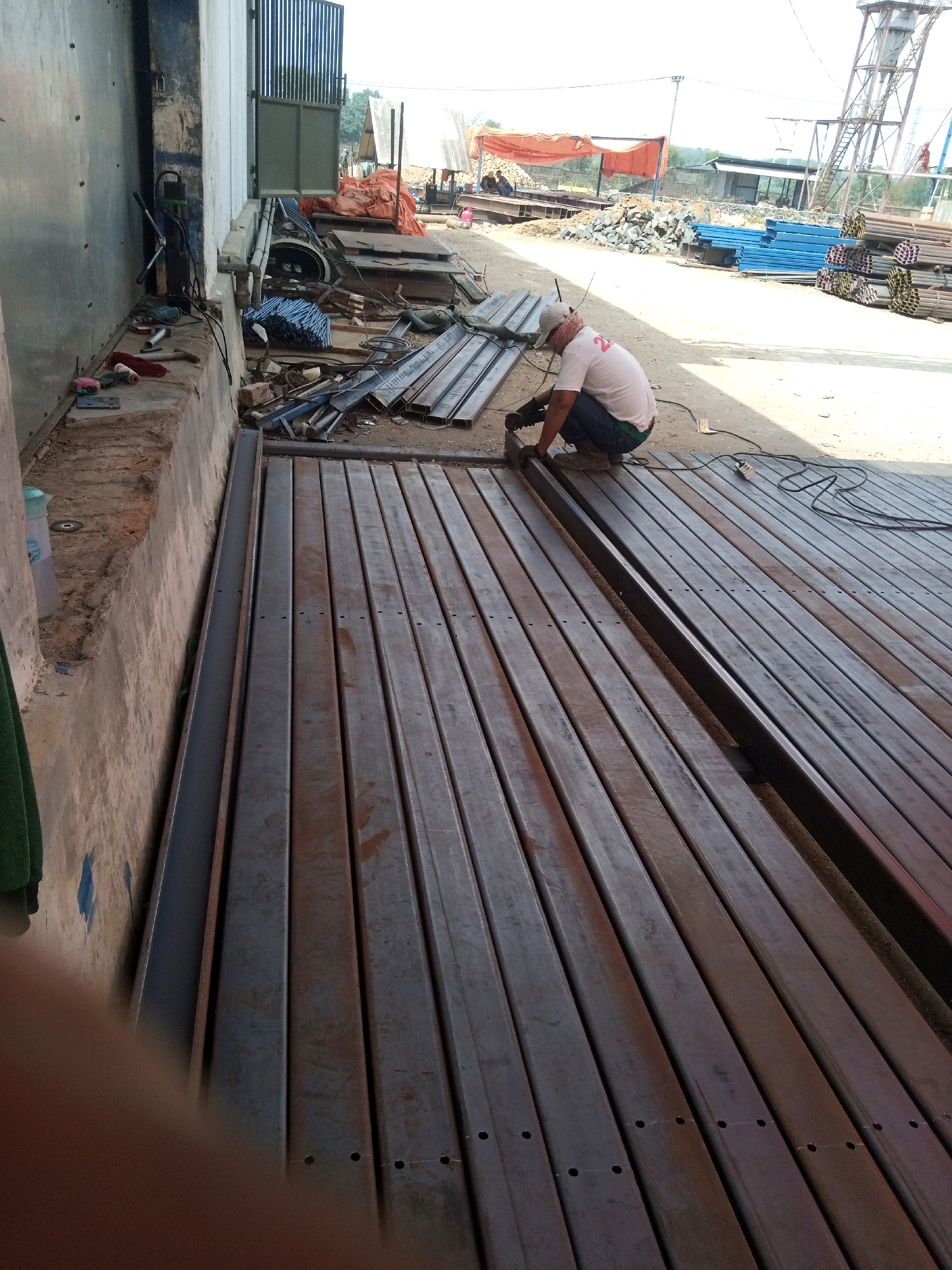 The image depicts a construction site in Teluk Dalem Ilir, Indonesia. In the foreground, a man wearing a white t-shirt, a baseball cap, and dark pants is crouched down, working on a metal structure. He appears to be welding or grinding metal with a tool in his right hand, and there are welding sparks visible. The man is surrounded by numerous steel beams, laid out in rows on the ground. Behind him, more construction materials are piled up, including stacks of blue pipes and orange tarps. In the background, there is a tall structure resembling a water tower or industrial equipment, along with some buildings and trees, suggesting a semi-urban or industrial area. The ground is dirt and gravel. The overall scene suggests active construction or fabrication work.