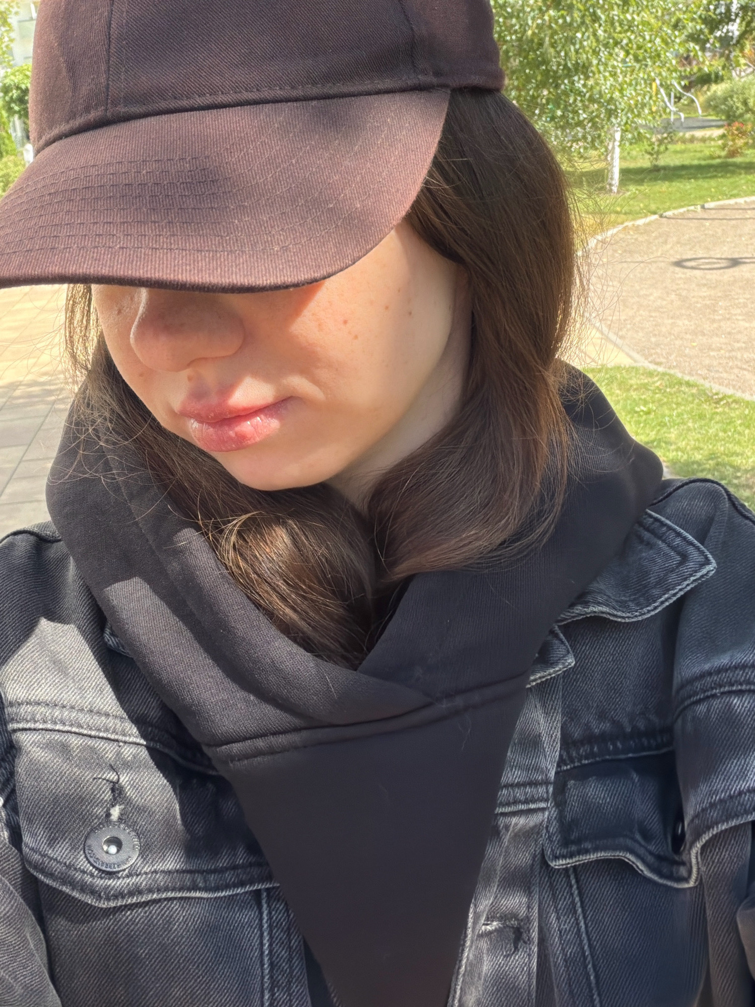 The image shows a close-up of a young person wearing a dark brown baseball cap, a black hoodie, and a dark denim jacket. The cap is tilted forward, obscuring the upper part of the face, but revealing the nose and lips. The person has long, dark brown hair and appears to be outdoors in a park or outdoor recreational area. The background shows a paved walkway, green grass, and trees, suggesting a sunny day. There is no indication of the location being Kyiv, Ukraine.