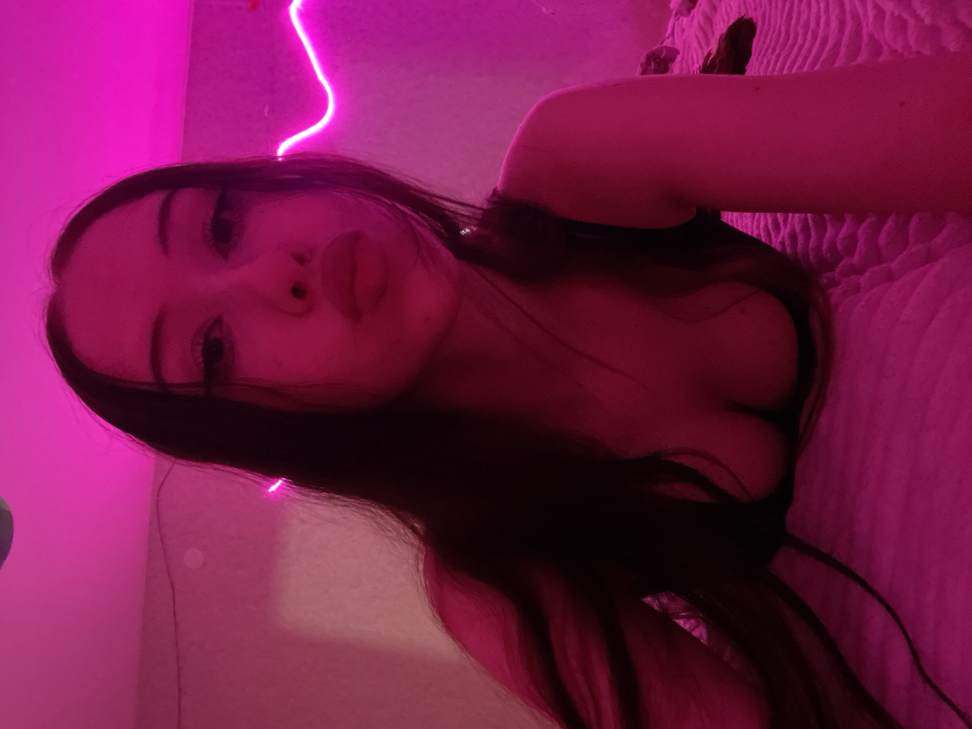 A young woman with long dark hair and visible cleavage is lying on a bed, illuminated by pink neon lights. The room is dimly lit, with the pink light casting a strong glow on her skin and the surrounding environment. Her face is turned towards the camera, with her lips slightly parted. The background shows a textured wall and a neon light fixture. There is no indication of the location in Votkinsk, Russia.