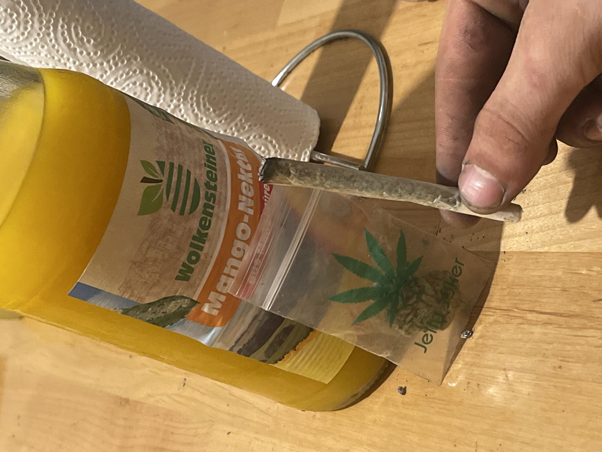 An overhead, close-up shot shows a person's right hand holding a marijuana cigarette, which is partially rolled. The cigarette is positioned in front of a plastic bag that contains marijuana bud. Behind the bag is a bottle of Wolkensteiner Mango-Nektar, a yellow beverage, and a white paper towel roll on a metallic stand. The scene is set on a wooden surface.