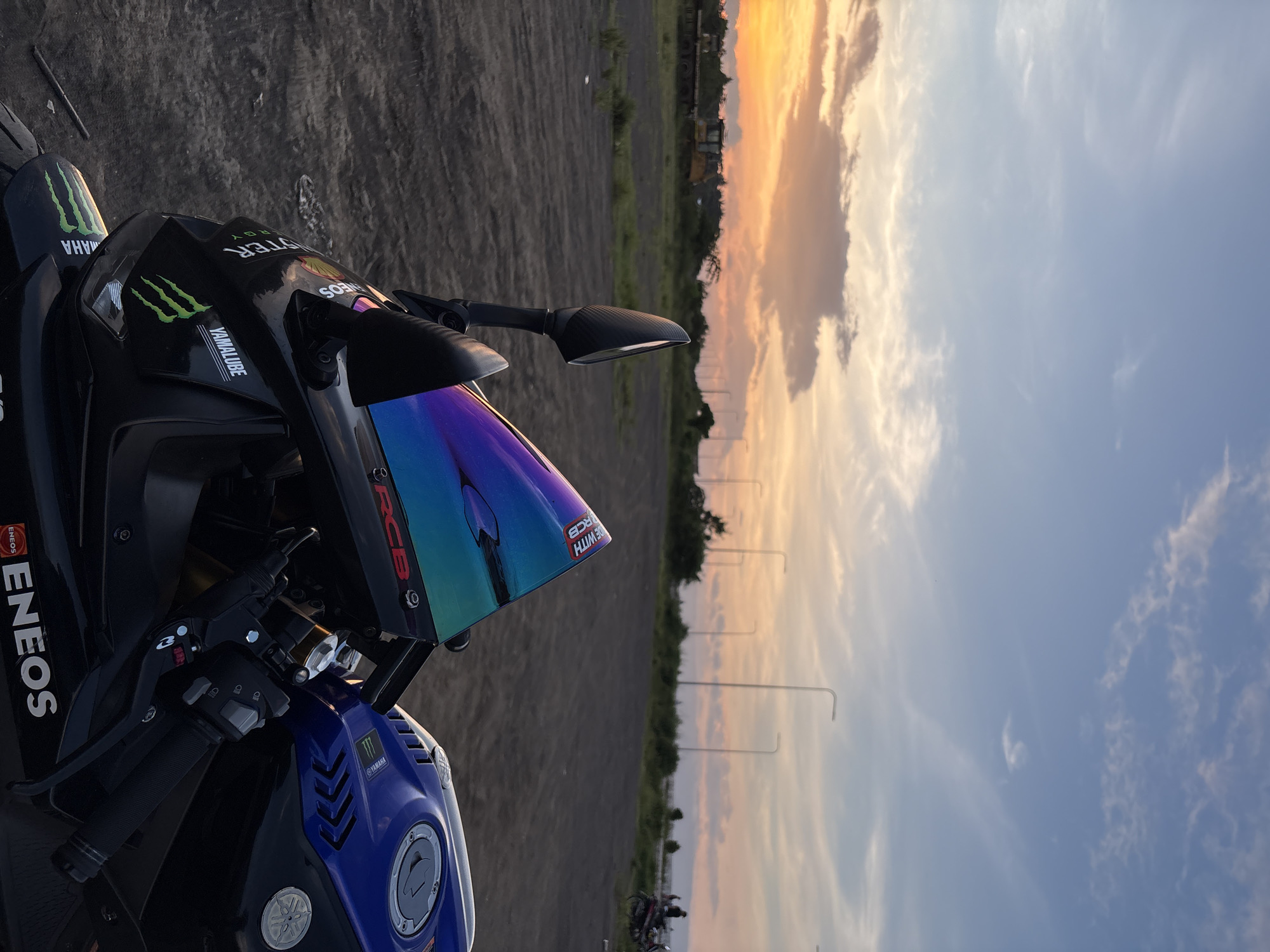 The image shows a black and blue Yamaha motorcycle parked on a dirt and gravel surface during sunset. The motorcycle has a colorful, iridescent windshield with "RCB" and "SMITH" branding. The fuel tank is blue and black with Yamaha and Monster Energy logos. The background depicts a flat, open area with some vegetation, utility poles with streetlights, and construction equipment in the distance. The sky is a gradient of blue, orange, and yellow, indicating sunset. There are a few distant figures and motorcycles visible on the right side of the frame. The setting appears to be an undeveloped or industrial area on the outskirts of a city.
This image may have been taken in Davao City, Philippines, specifically in areas like the coastal road or near industrial zones where such open, undeveloped spaces with views of the sunset are common. For example, areas along the Francisco Bangoy International Airport road or the coastal areas of Toril District could present similar landscapes.