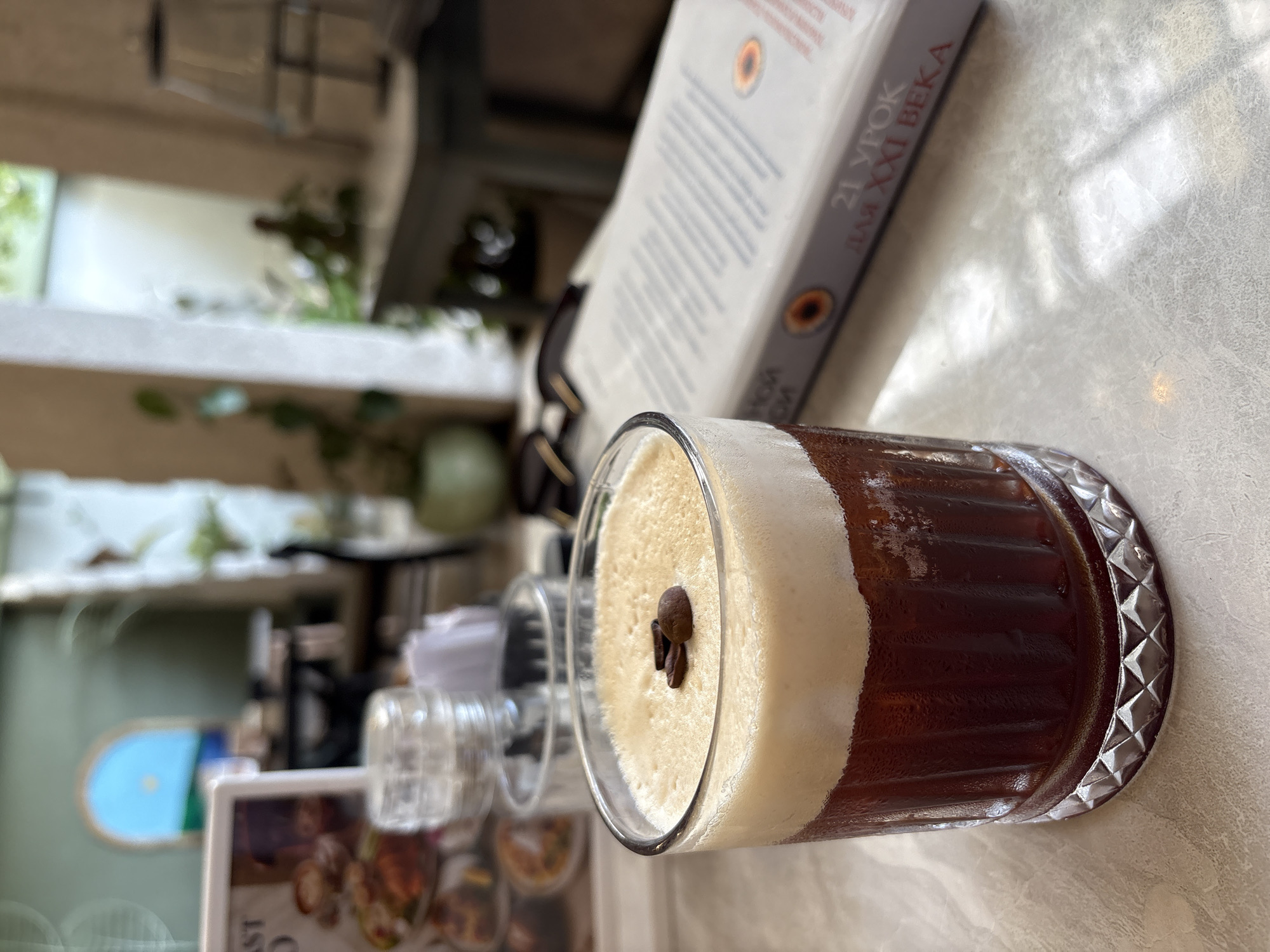 An espresso drink with a thick layer of foam topped with coffee beans sits on a table. The drink is in a clear glass with a decorative pattern around the bottom. In the background, a book with Cyrillic text titled "21 УРОК ДЛЯ XXI ВЕКА" is visible. The scene appears to be indoors, possibly a cafe or restaurant, with a blurred background showing plants and other tables. The lighting suggests it could be daytime.