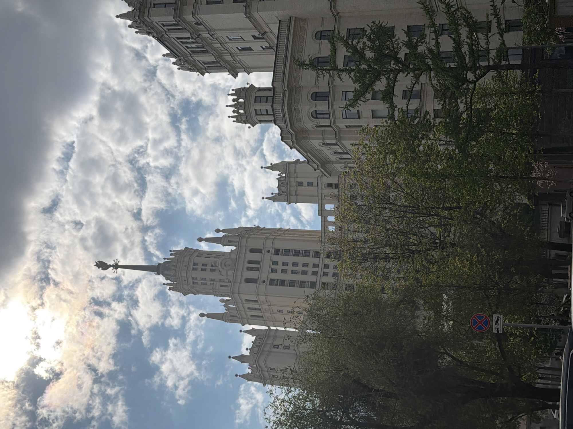 The image shows a tall, ornate building with a spire against a cloudy sky. The building is light-colored and features multiple tiers with decorative architectural details. Trees with green leaves partially obscure the building at the lower portion of the frame. A traffic sign with a red and blue "X" is visible on the right side of the image, partially hidden by tree branches. The sun is behind the clouds on the left side of the image, creating a bright glow. The image appears to be taken from a low angle, looking up at the building and sky. I cannot make claims about the location beyond what is provided in the image.