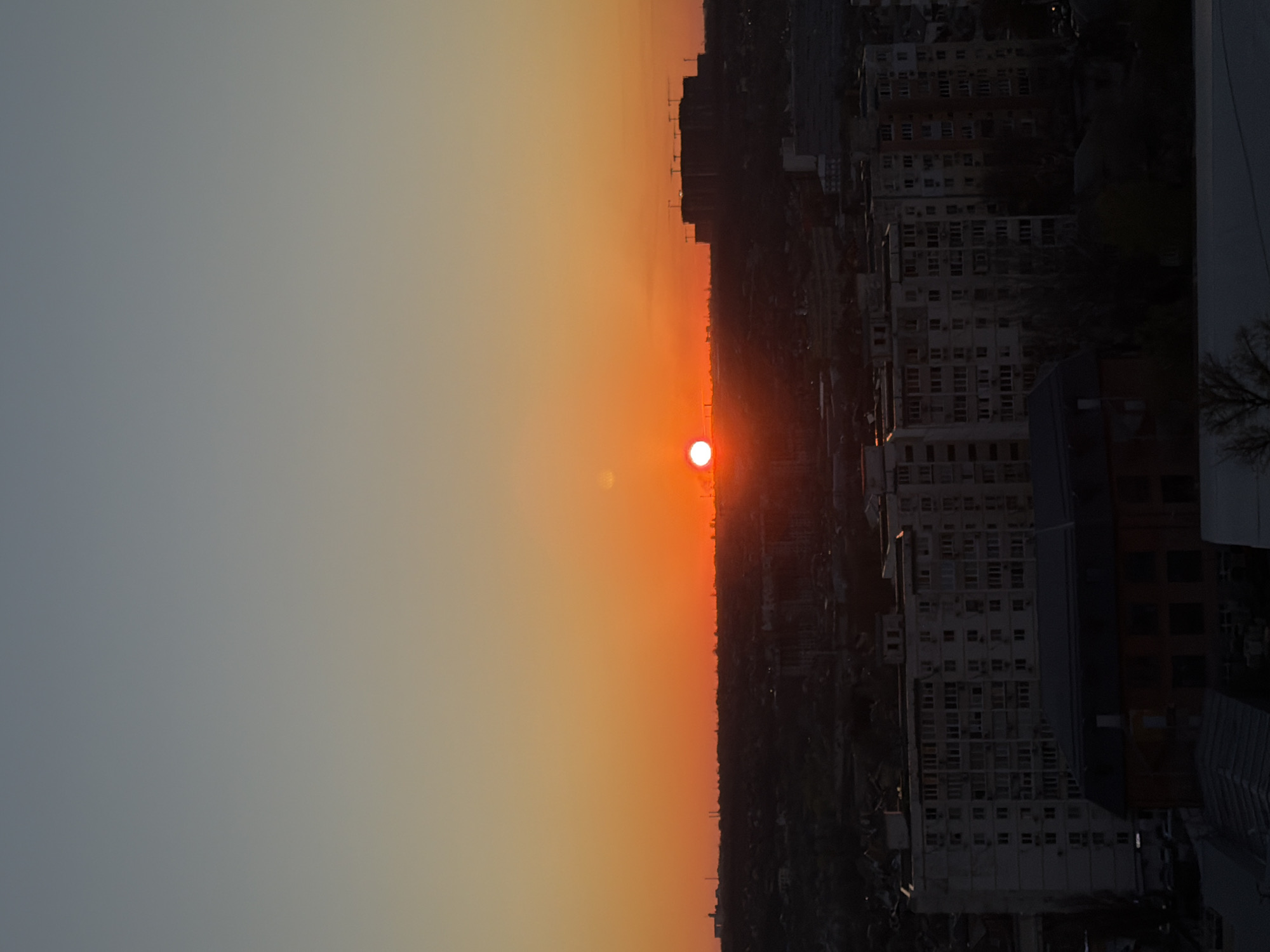 The image depicts a sunset over a city, likely Rostov-on-Don, Russia. A bright, orange sun sits on the horizon line, casting a warm glow across the sky, which transitions from a light gray-blue to a deep orange near the horizon. The silhouetted cityscape comprises numerous apartment buildings with rows of windows. The buildings are predominantly white, with some dark sections. The overall scene evokes a sense of urban life against the backdrop of a natural phenomenon.