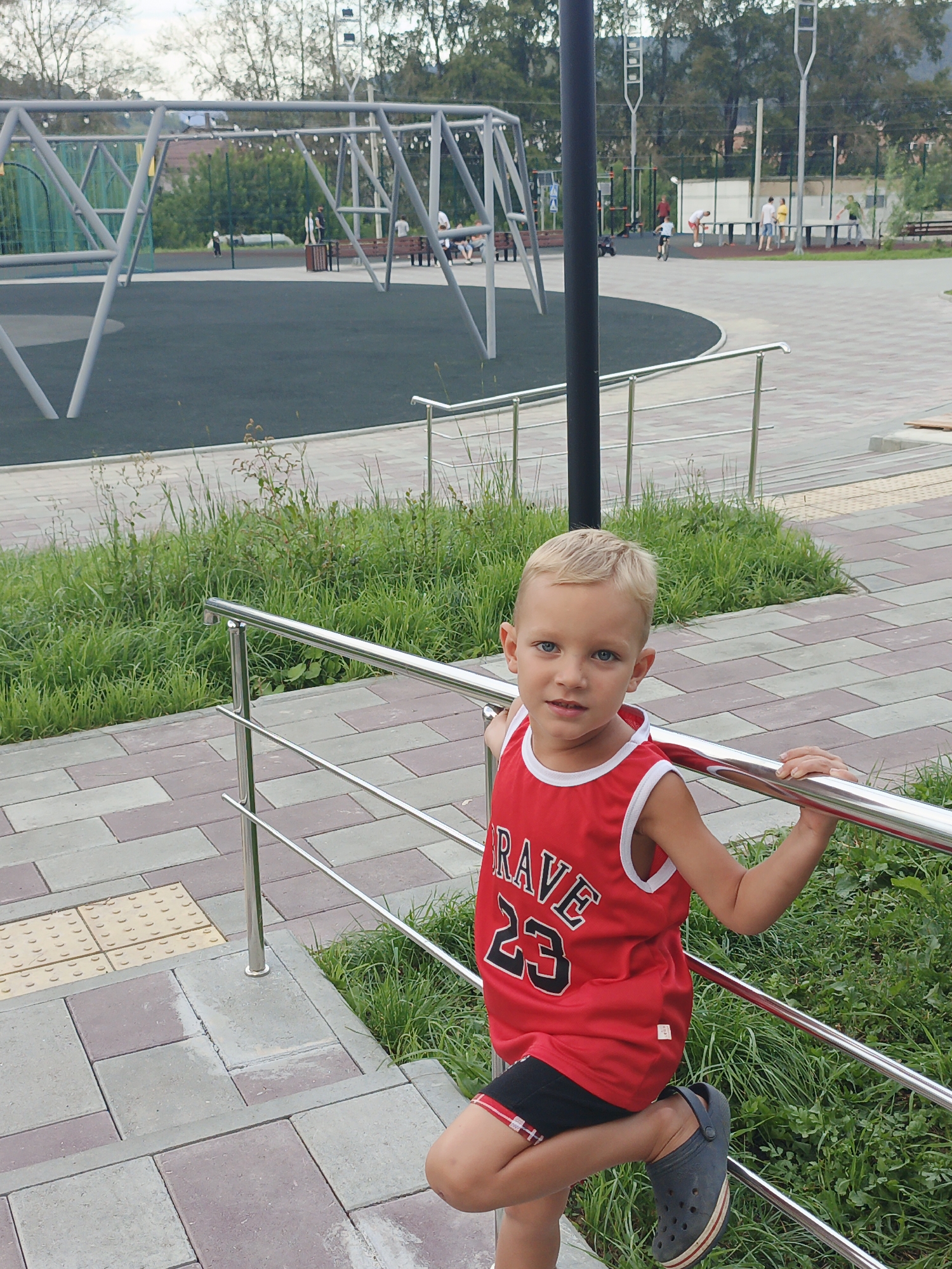 A young boy with blonde hair and blue eyes, wearing a red basketball-style jersey with "RAVE 23" printed on it and black shorts, poses for a photo. He is holding onto a silver railing at a playground. The playground has a rubberized surface and swings in the background. The boy is on a tiled pathway. The scene appears to be in Zlatoust, Russia, based on the context provided.