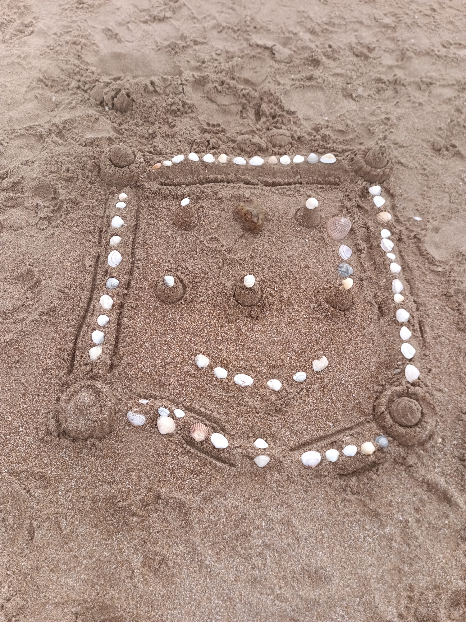 The image shows a sandcastle on the beach in Houlgate, France, decorated to resemble a face. The sandcastle is roughly square-shaped with rounded corners, and features two small sand mounds topped with shells for eyes, and a line of shells forming a smile. Two small, conical sand structures are positioned above the eyes, and a larger, round object, possibly a stone or a larger shell, is placed between them. The perimeter of the sandcastle is decorated with small shells. The background is composed of damp sand, showing footprints and textures typical of a beach environment.