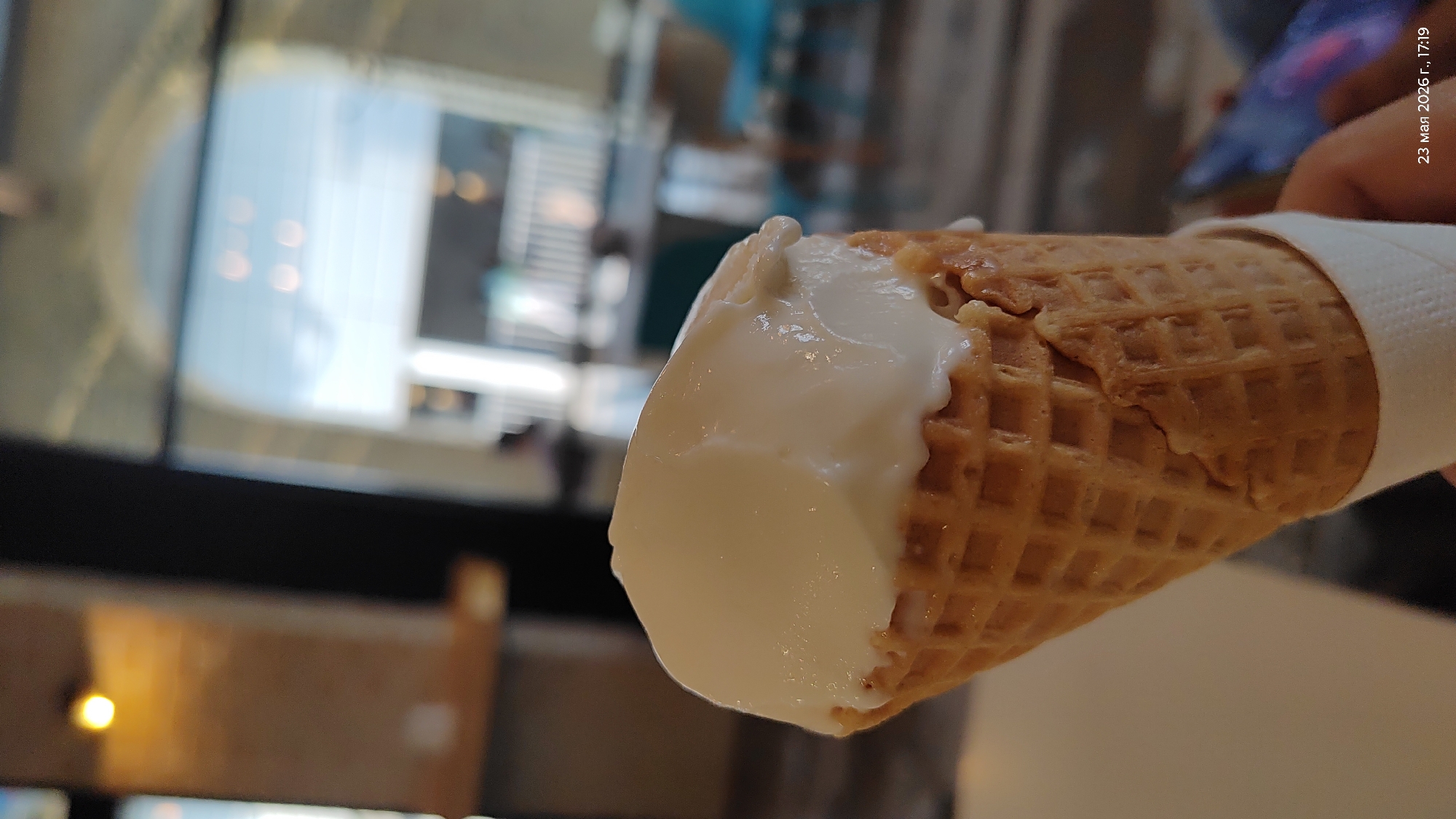 The image shows an ice cream cone with vanilla ice cream partially eaten, held in someone's hand. A white napkin is wrapped around the cone. The background appears to be a blurred interior with a large glass window showing a cityscape. The timestamp on the image reads "23 мая 2026 г., 17:19". The details are not precise enough to define that this image was taken in Almaty.