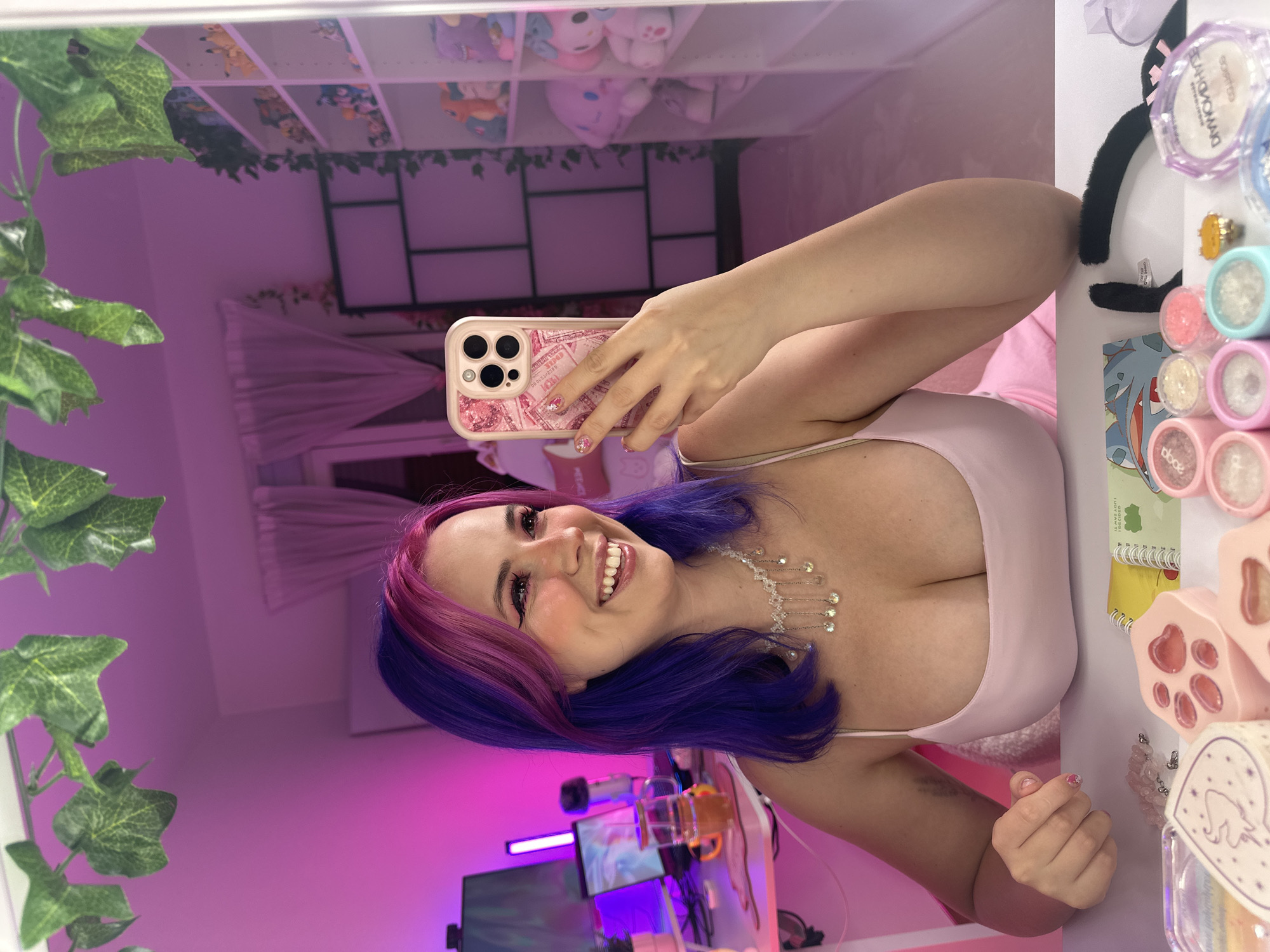A woman with purple and pink dyed hair, wearing a light pink camisole and a sparkling necklace, smiles as she takes a selfie in a mirror. Her arm is raised to hold a pink, glittery phone case. The background, reflected in the mirror, shows shelves filled with stuffed animals, a window with sheer curtains, and a desk with a computer monitor, microphone, and a glass of amber liquid. Green ivy vines are draped above the mirror. In the foreground on a white surface, there are various cosmetic items, including small pots of glitter, a ring with a yellow gem, and a black cat ear headband. The room is illuminated with soft pink lighting. There is no discernible information or context within the image to suggest a location in Oviedo, Spain.